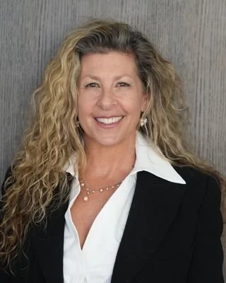 A smiling woman with curly blonde hair, wearing a black blazer over a white shirt, standing against a gray background.