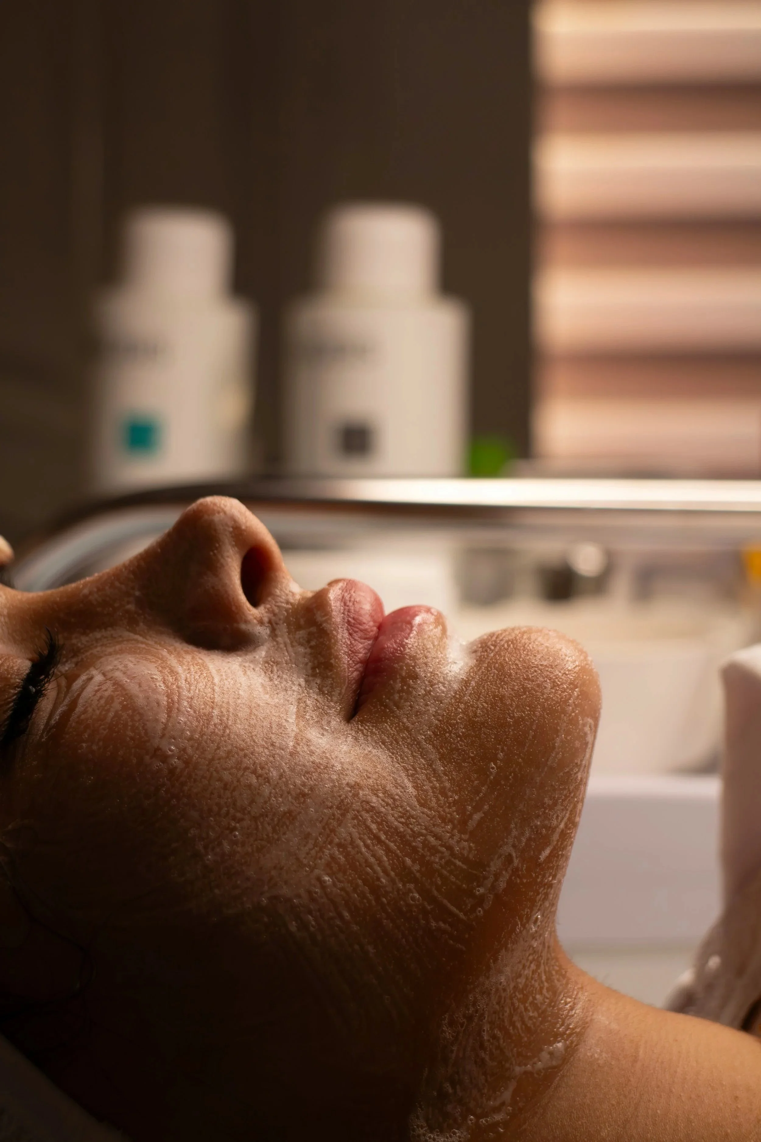 Close-up of a person lying face-up with their eyes closed, appearing relaxed, in a spa or wellness setting, with bottles of skincare products in the background.