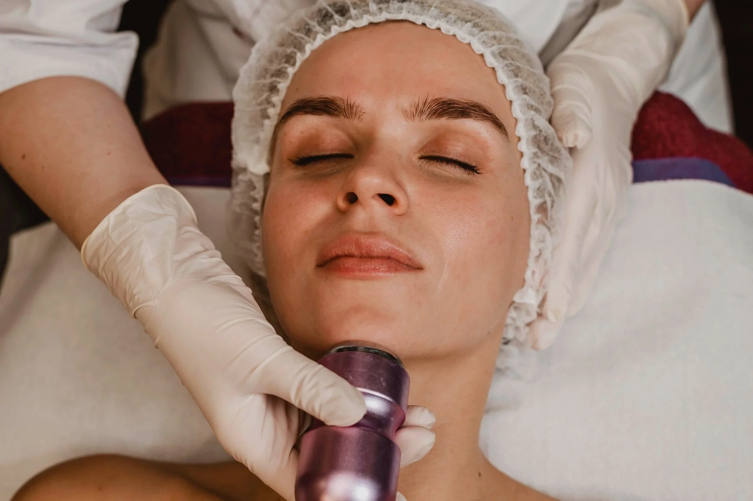 A woman lying on a medical bed with eyes closed, wearing a hair cap, receiving cosmetic treatment as a gloved hand holds a device near her face.