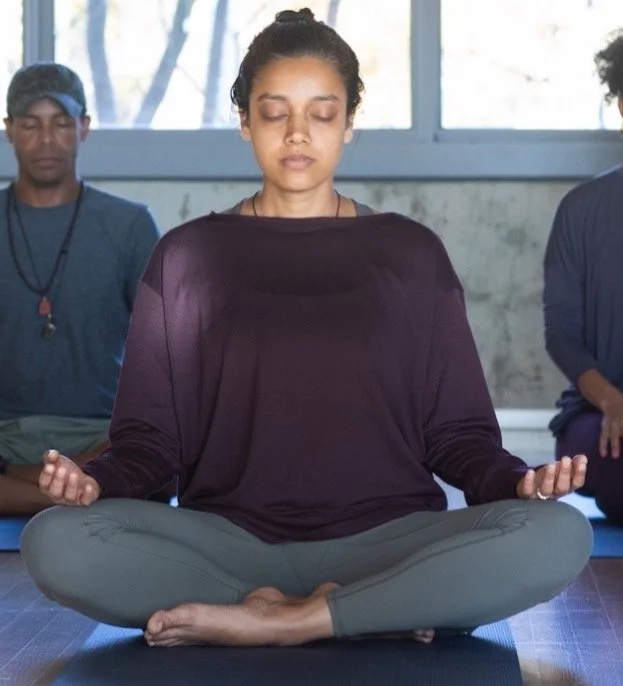 A woman practicing meditation in a cross-legged seated position with eyes closed, wearing a loose dark purple top and gray leggings, in a peaceful indoor setting with others in the background.