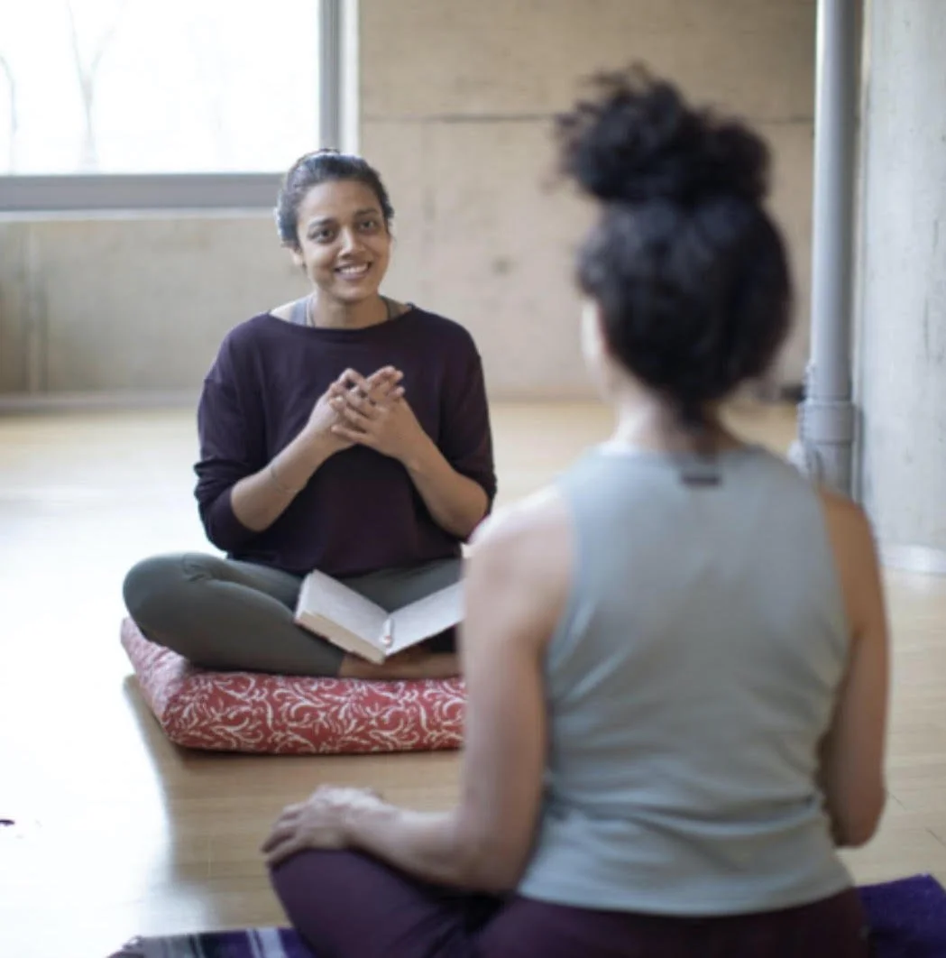 Two women practicing yoga or meditation in a bright studio with a window. One woman is sitting cross-legged on a cushion, smiling, with a book open on her lap, while the other woman is sitting with her back to the camera.