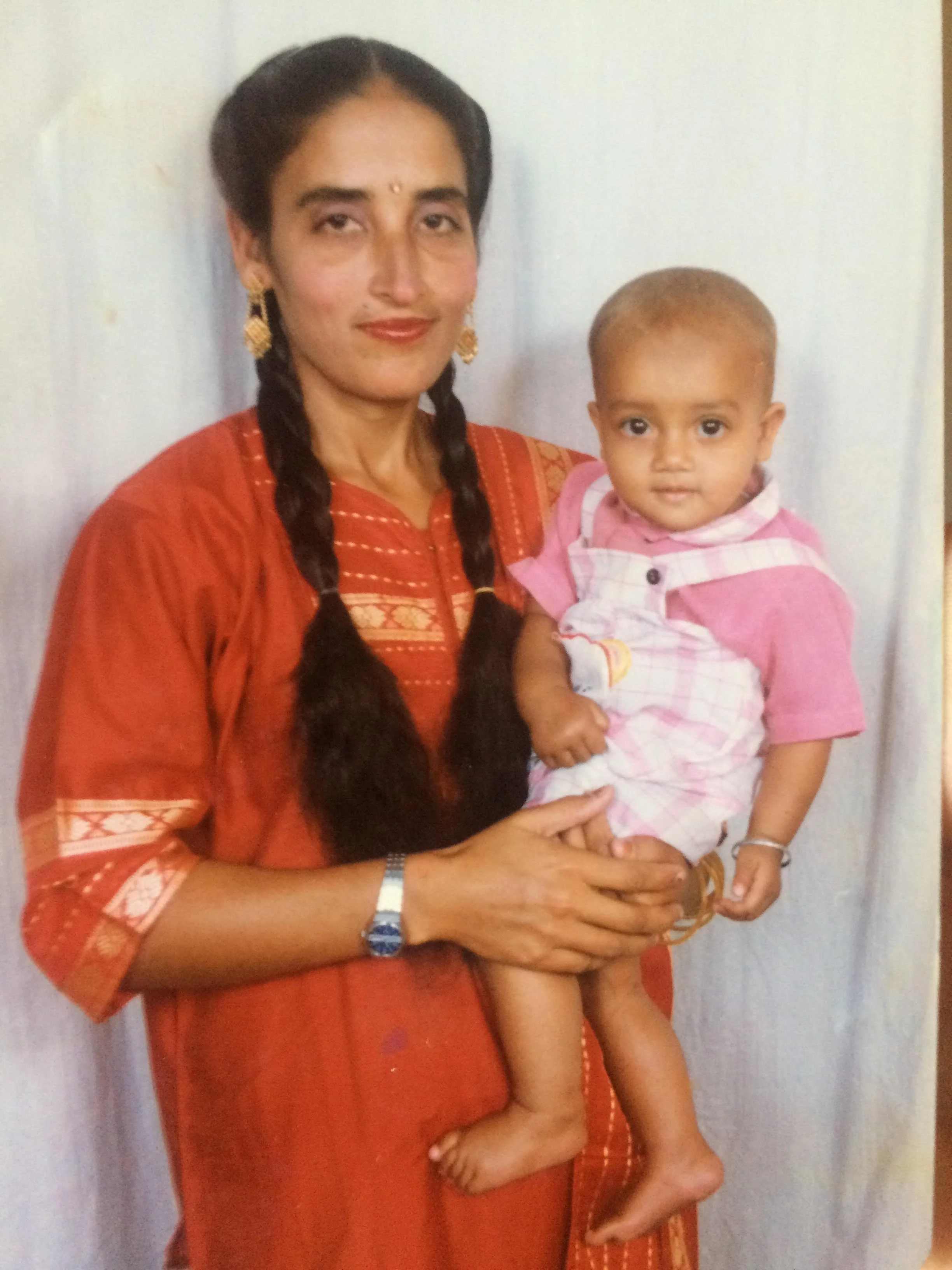 A woman with long dark hair braided into two pigtails, wearing traditional Indian jewelry and orange ethnic attire, holding a young child with a shaved head, big eyes, and wearing a pink checkered shirt, standing against a plain white fabric background.