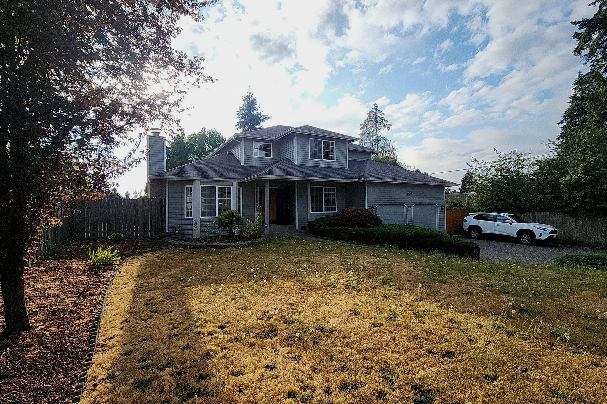 A two-story gray house with multiple windows, a covered front porch, and a two-car garage. A white SUV is parked in the driveway. The yard has some grass, shrubs, and small plants. Tall trees and a wooden fence surround the property, with a driveway on the right side. The sky is partly cloudy with some visible clouds.