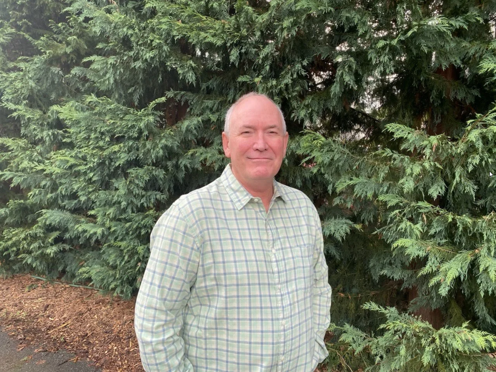 A middle-aged man with light skin, short gray hair, wearing a light green and white plaid shirt, standing outdoors in front of green evergreen trees.