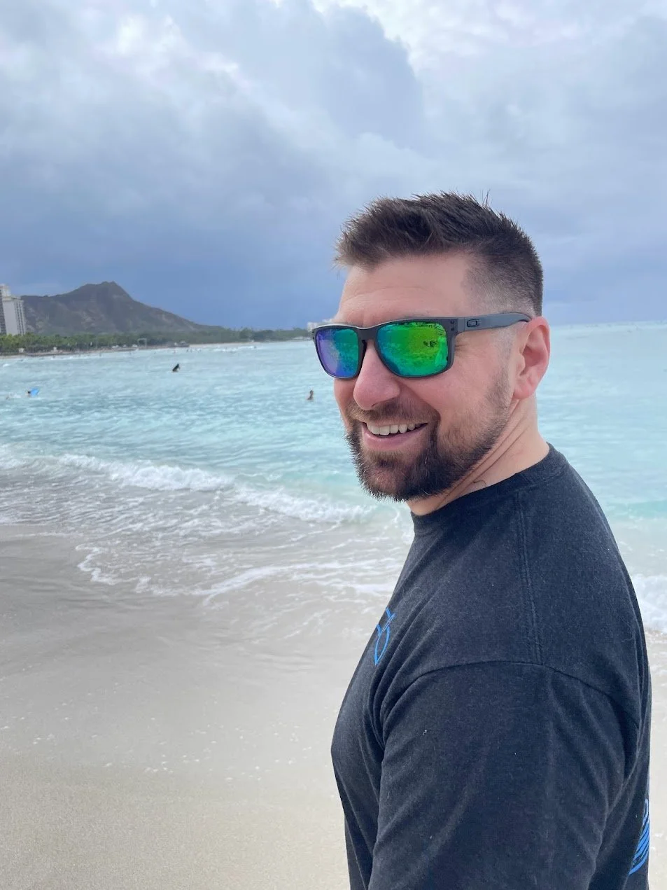 Smiling man wearing sunglasses on a beach with ocean waves and cloudy sky in the background.