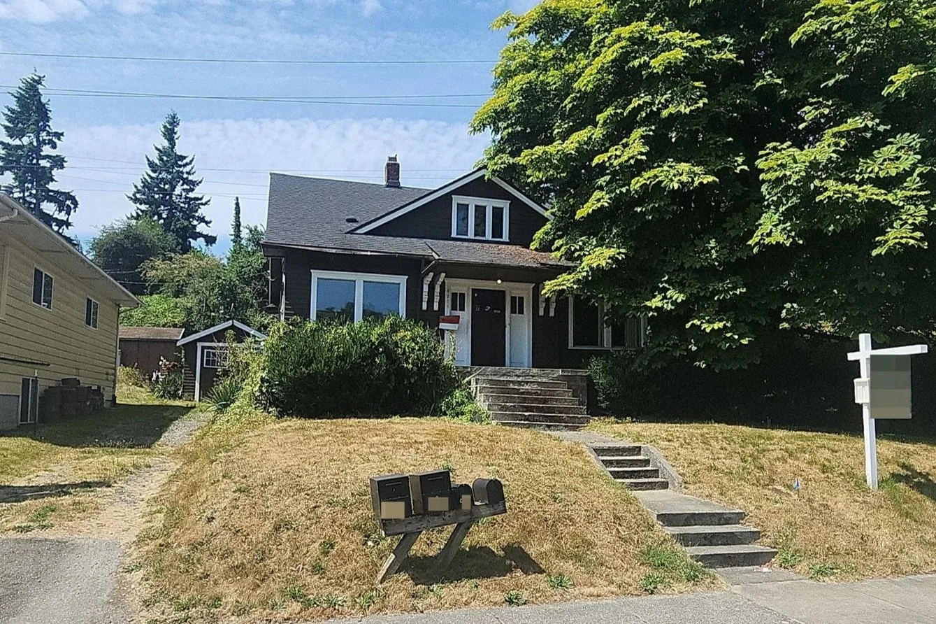 Front view of a black house with stairs leading up to the entrance, surrounded by green trees and shrubs, with a mailbox and mailboxes on the lawn.