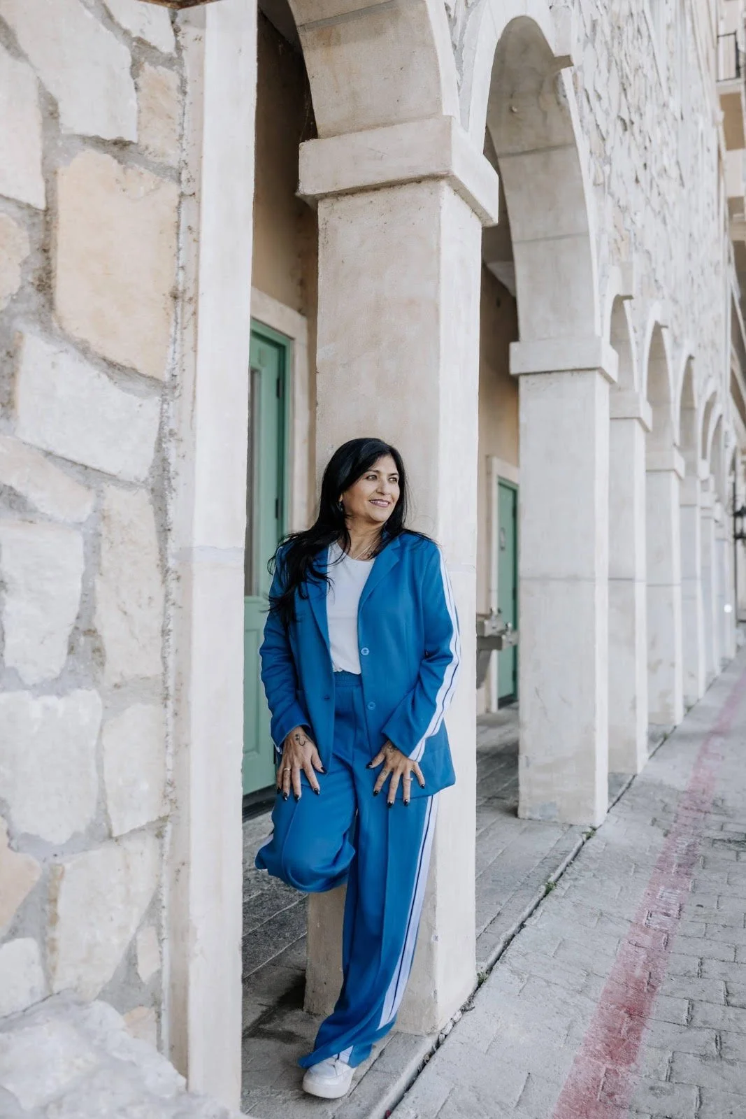 A woman with black hair wearing a blue jacket and matching blue pants with white stripes, standing in front of stone arches on a sidewalk.