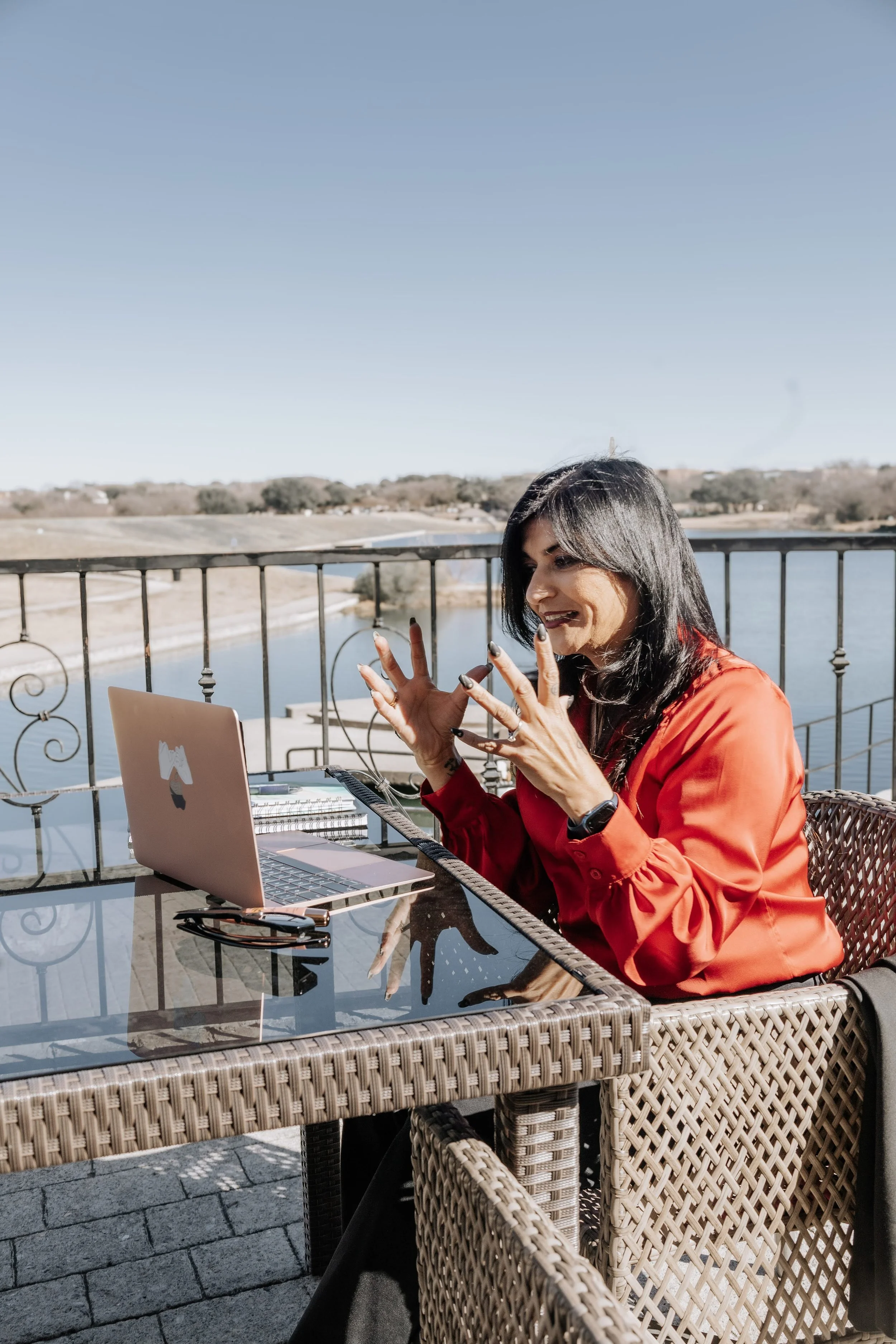 Woman with black hair, wearing a red blouse, sitting at a wicker outdoor table with a laptop, smiling and making gestures with her hands, overlooking a river and landscape under a clear blue sky.