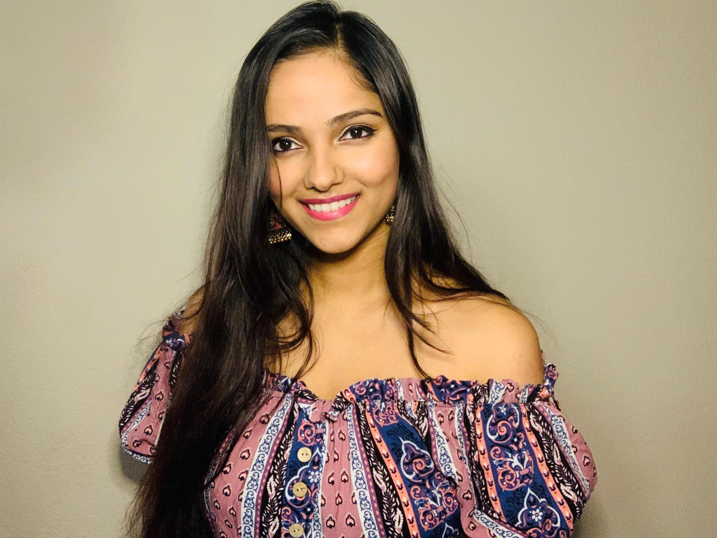 A young woman with long dark hair, wearing a colorful off-shoulder dress with a paisley pattern, smiling against a plain beige wall.