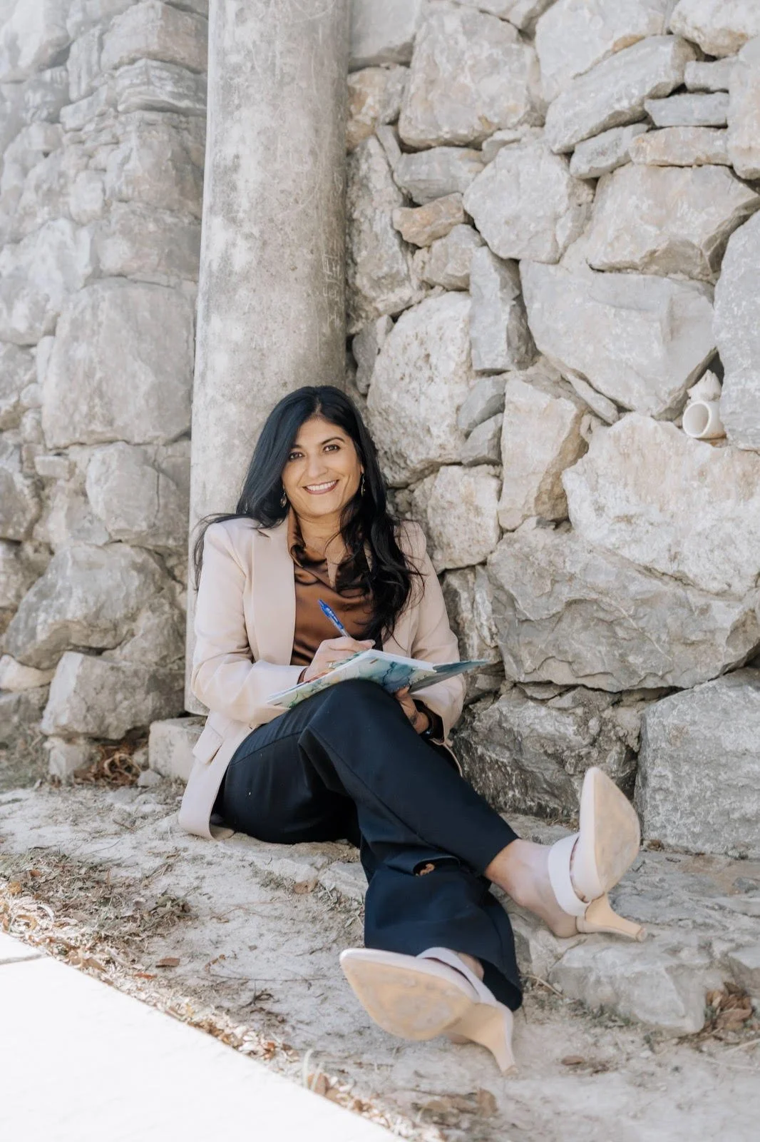 A smiling woman with dark hair, wearing a beige blazer, brown top, and black pants, sitting on the ground and writing on a notepad with a pen, leaning against a stone wall.