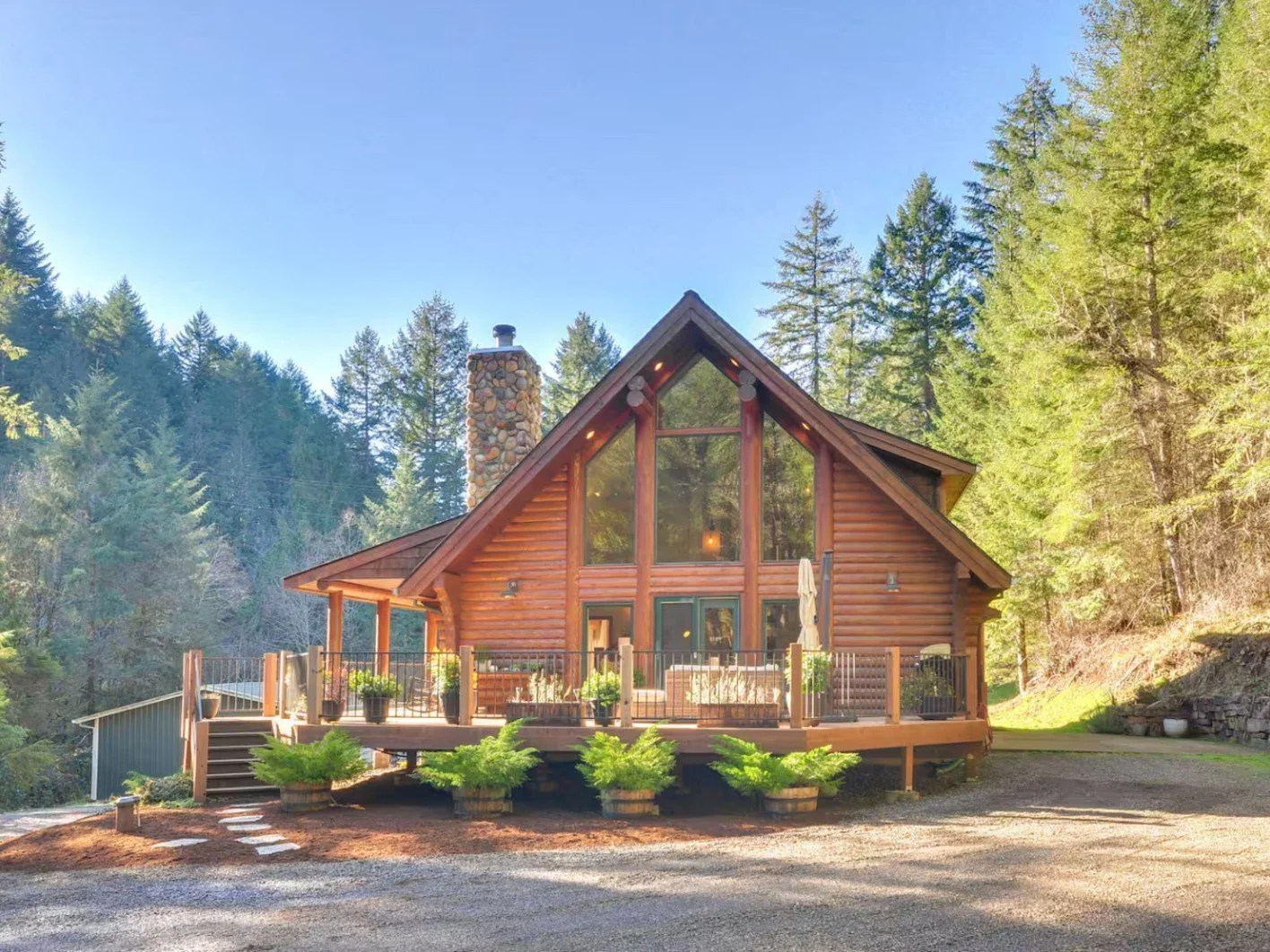 A beautiful log cabin house with large front windows, a stone chimney, and a spacious wooden deck surrounded by green trees and nature