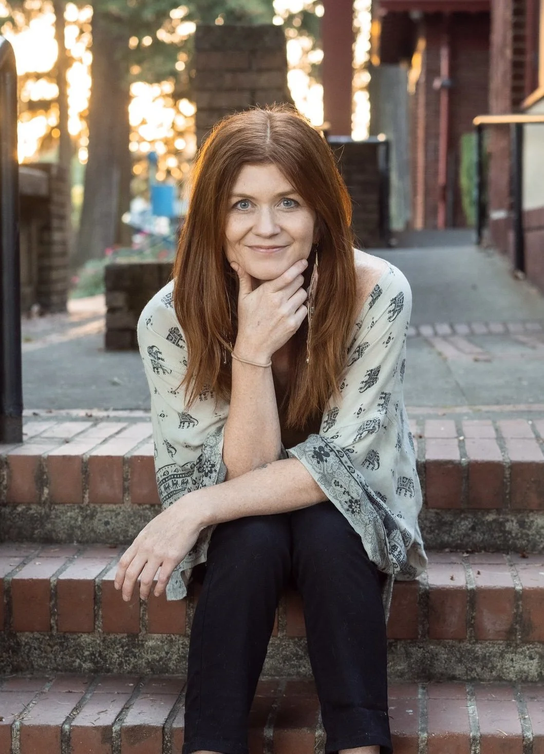 A woman with red hair sitting on brick steps outdoors, wearing a light-colored patterned top and black pants, resting her chin on her hand, smiling at the camera.