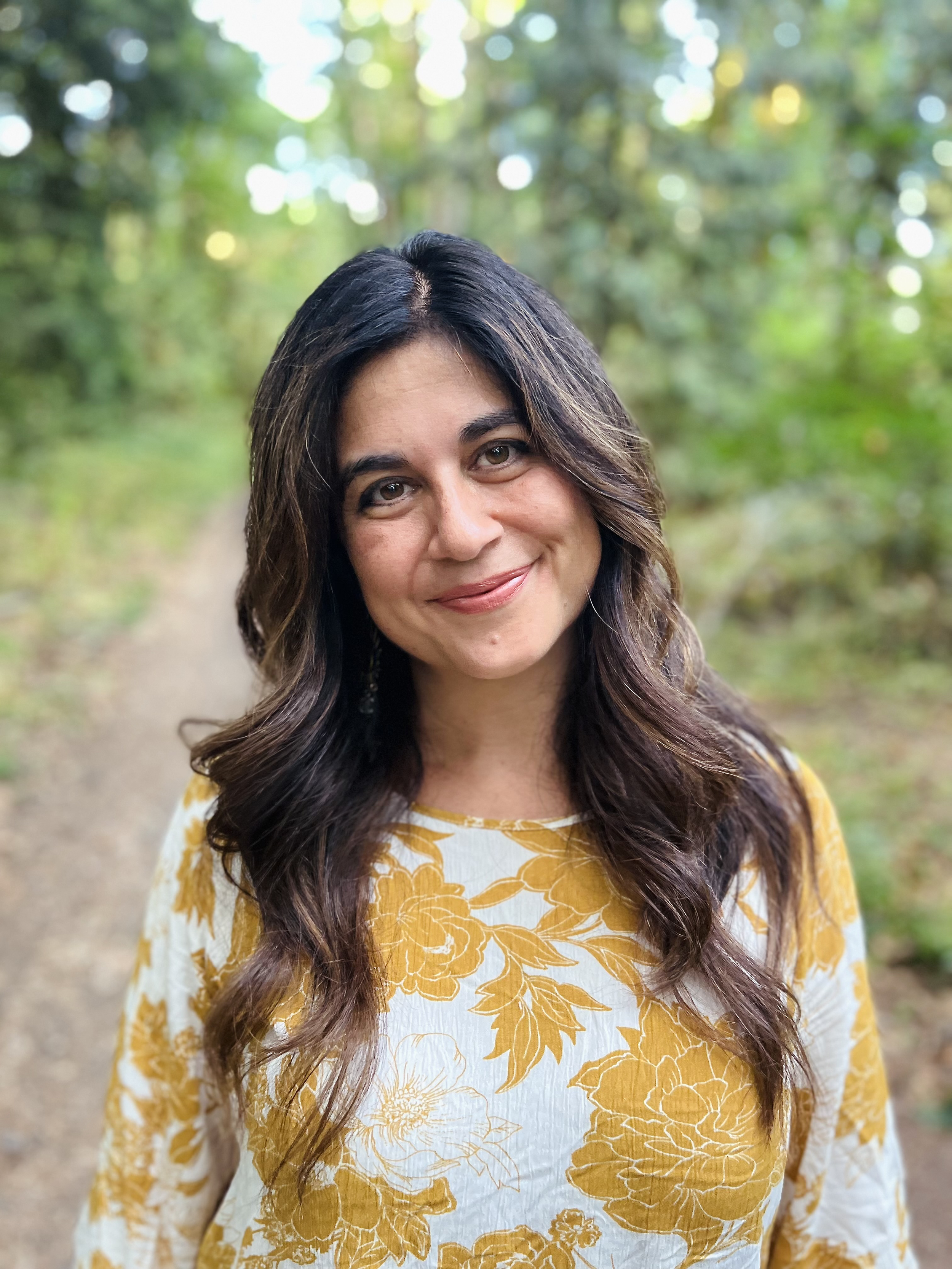 A woman with long, dark hair and brown eyes smiling outdoors in a natural setting with green trees and a dirt path in the background, wearing a yellow and white floral patterned top.