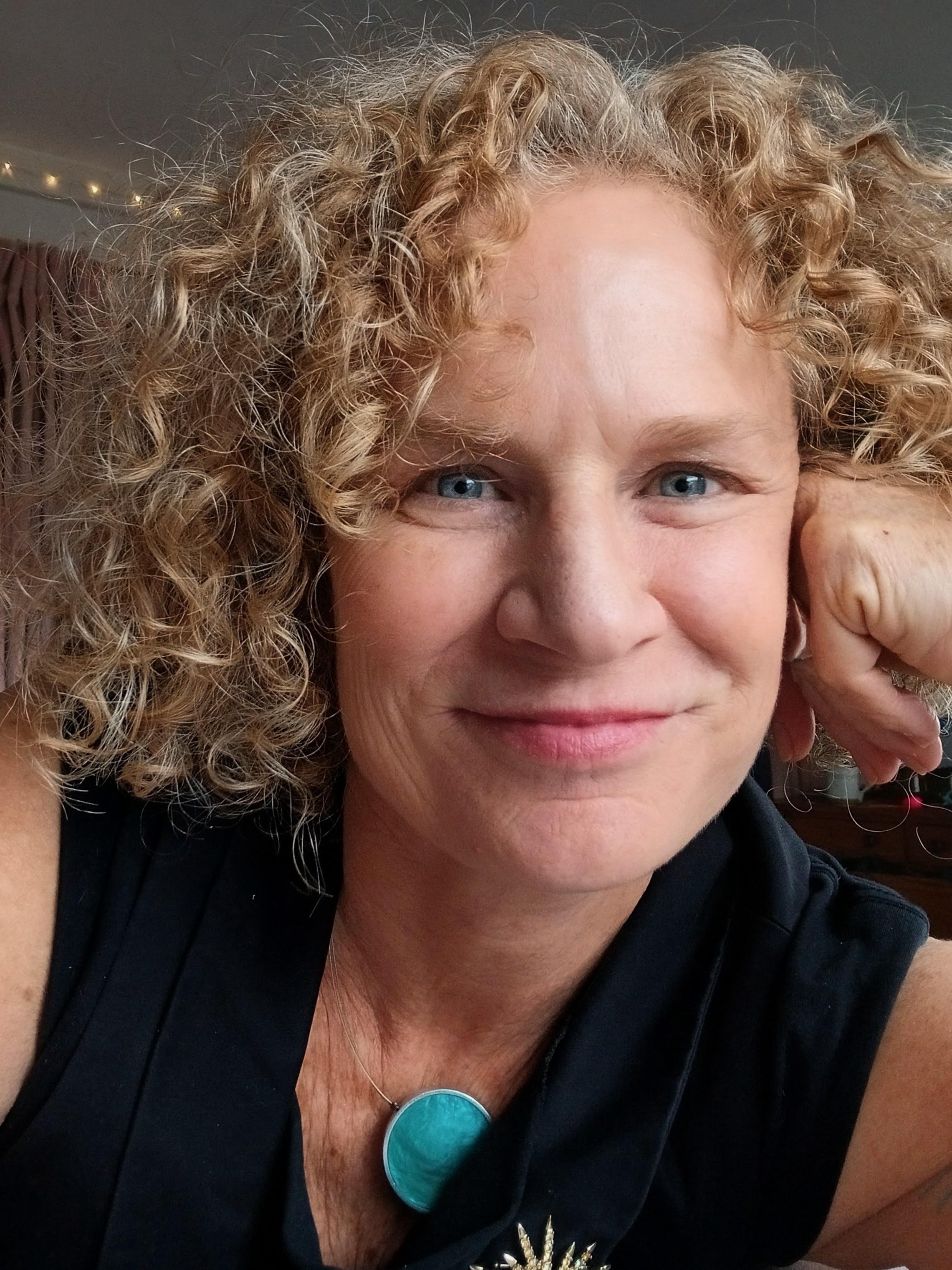 A woman with blonde curly hair, blue eyes, and light skin is smiling and resting her head on her hand. She is wearing a black top, a turquoise pendant necklace, and a ring on her finger.