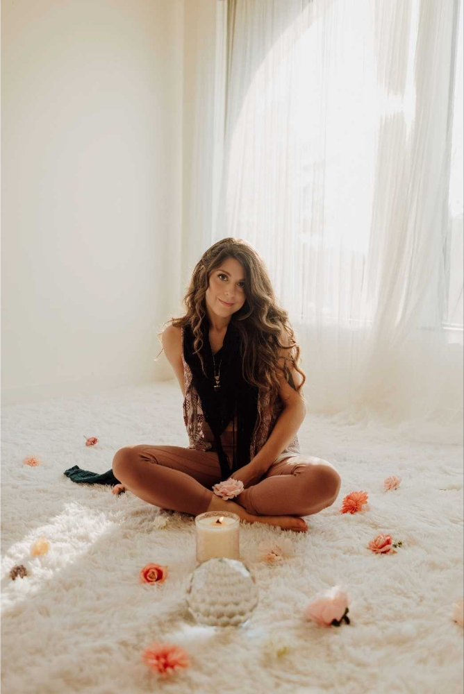 A woman sitting cross-legged on a white fluffy rug in a softly lit room with sheer curtains, surrounded by scattered flowers, with a candle and a decorative object in front of her.