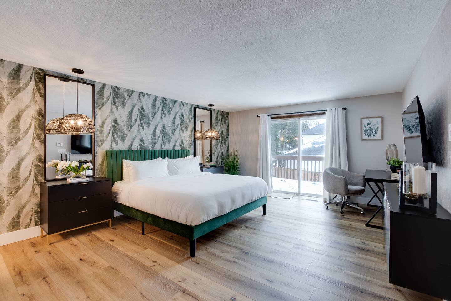 Modern hotel room with a large bed, green upholstered headboard, and sliding glass door leading to a balcony with snow outside. Decor includes tropical leaf wallpaper, black furniture, and a workspace with a chair and TV.
