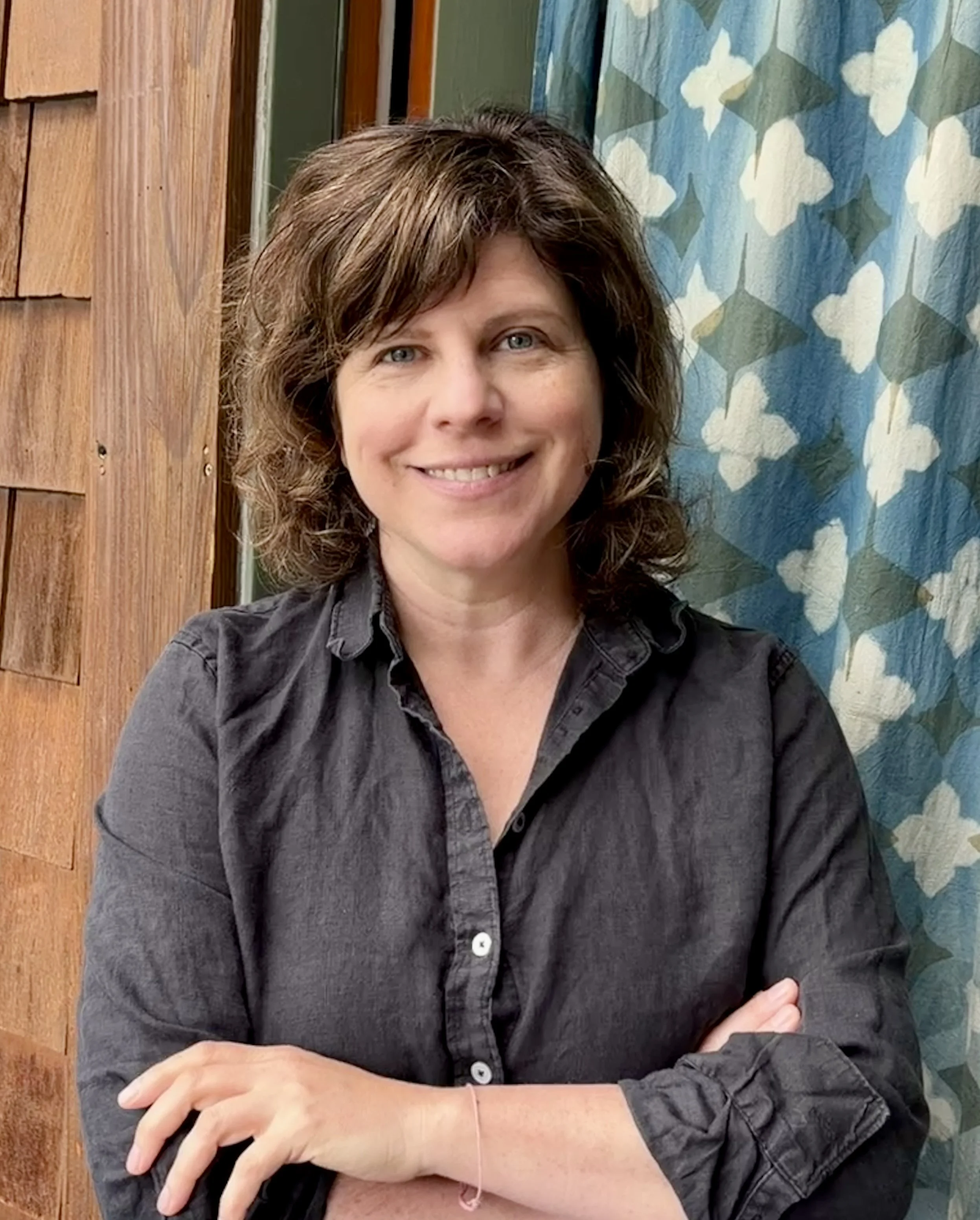 A woman with short wavy brown hair and blue eyes, smiling and sitting with her arms crossed, wearing a dark gray button-up shirt, in front of a wooden wall and patterned curtain.
