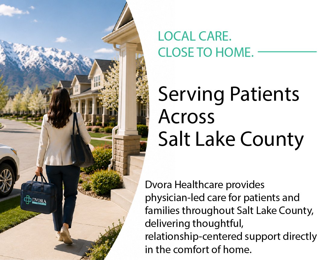 A woman in a beige blazer, navy pants, and beige shoes walking on a sidewalk in a suburban neighborhood with houses, snow-capped mountains, and cherry blossom trees in the background. She is carrying a black tote and a black bag with a healthcare provider logo.
