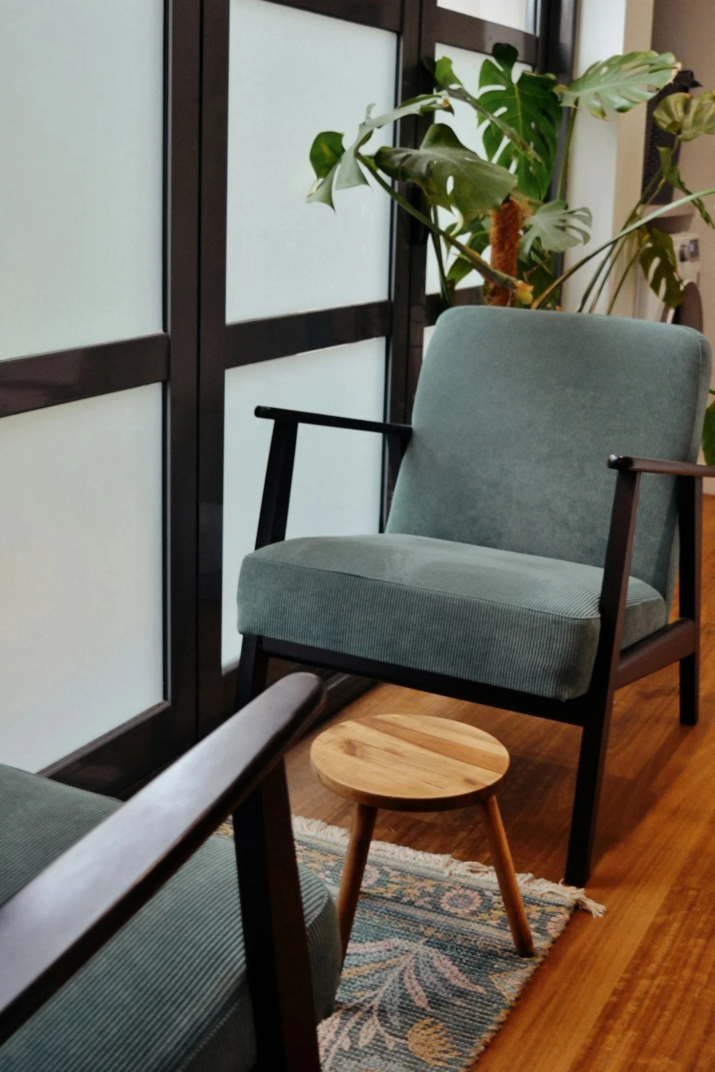 A cozy reading nook or sitting area with a green upholstered armchair, a small round wooden side table, a patterned rug, and a large leafy houseplant in the background.