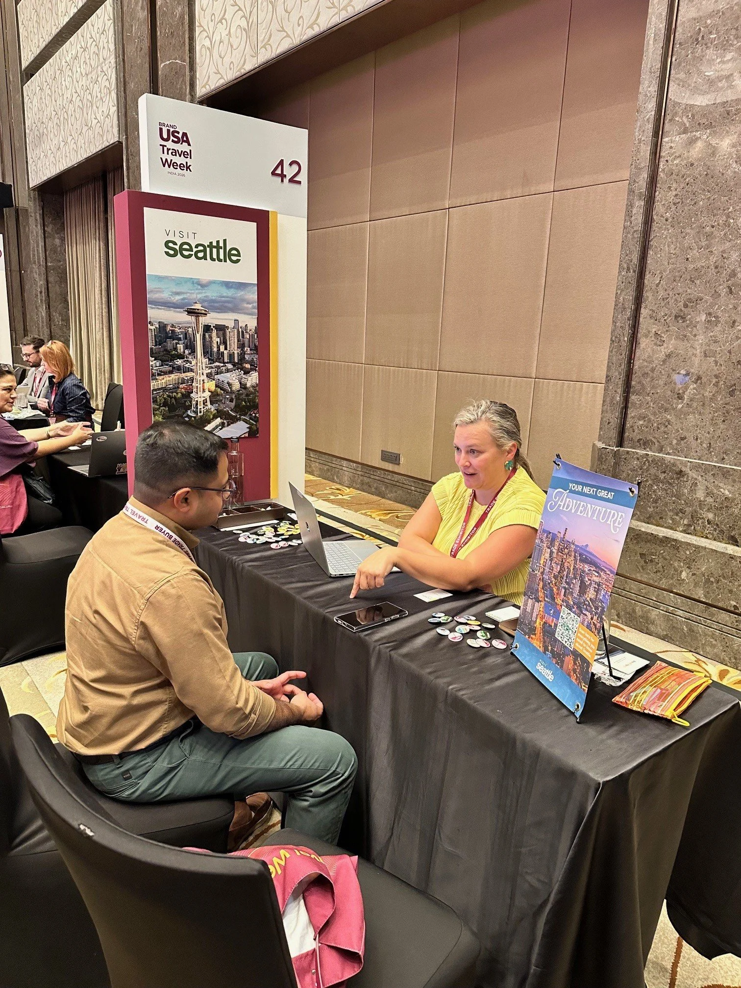 People sitting at a table at a travel event, with tables displaying promotional materials for Seattle and travel-themed brochures, in a hotel conference hall.