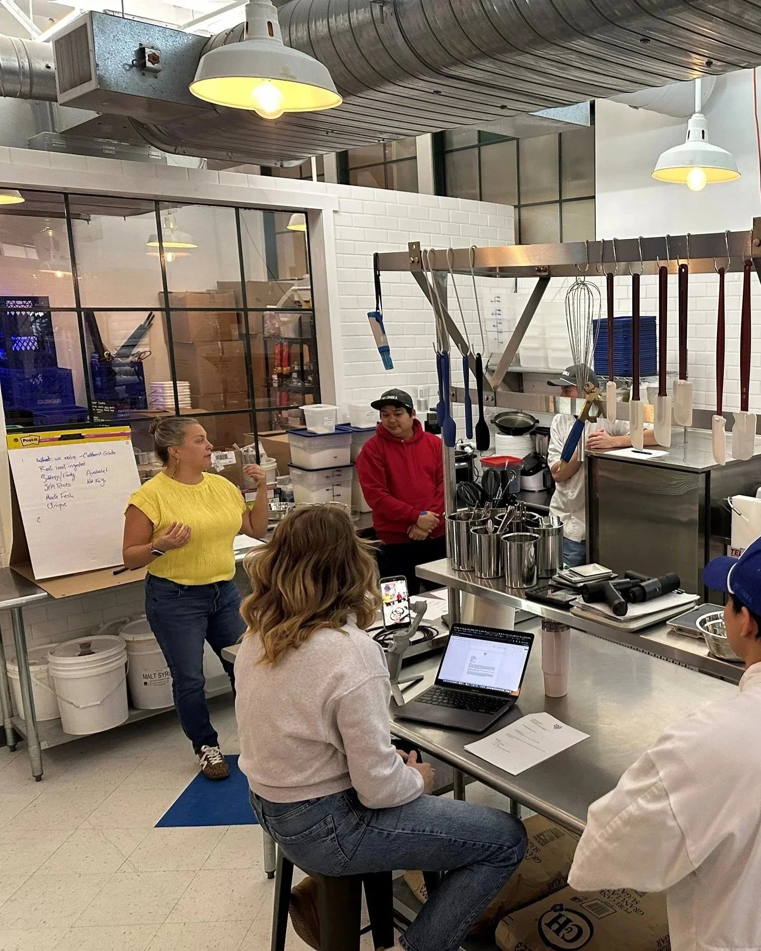 A woman in a yellow shirt is speaking to a group of people in a kitchen or industrial setting. Several individuals are seated at tables with laptops, while others stand near kitchen equipment. The background shows stainless steel containers, a white brick wall, and kitchen tools hanging from a rack.