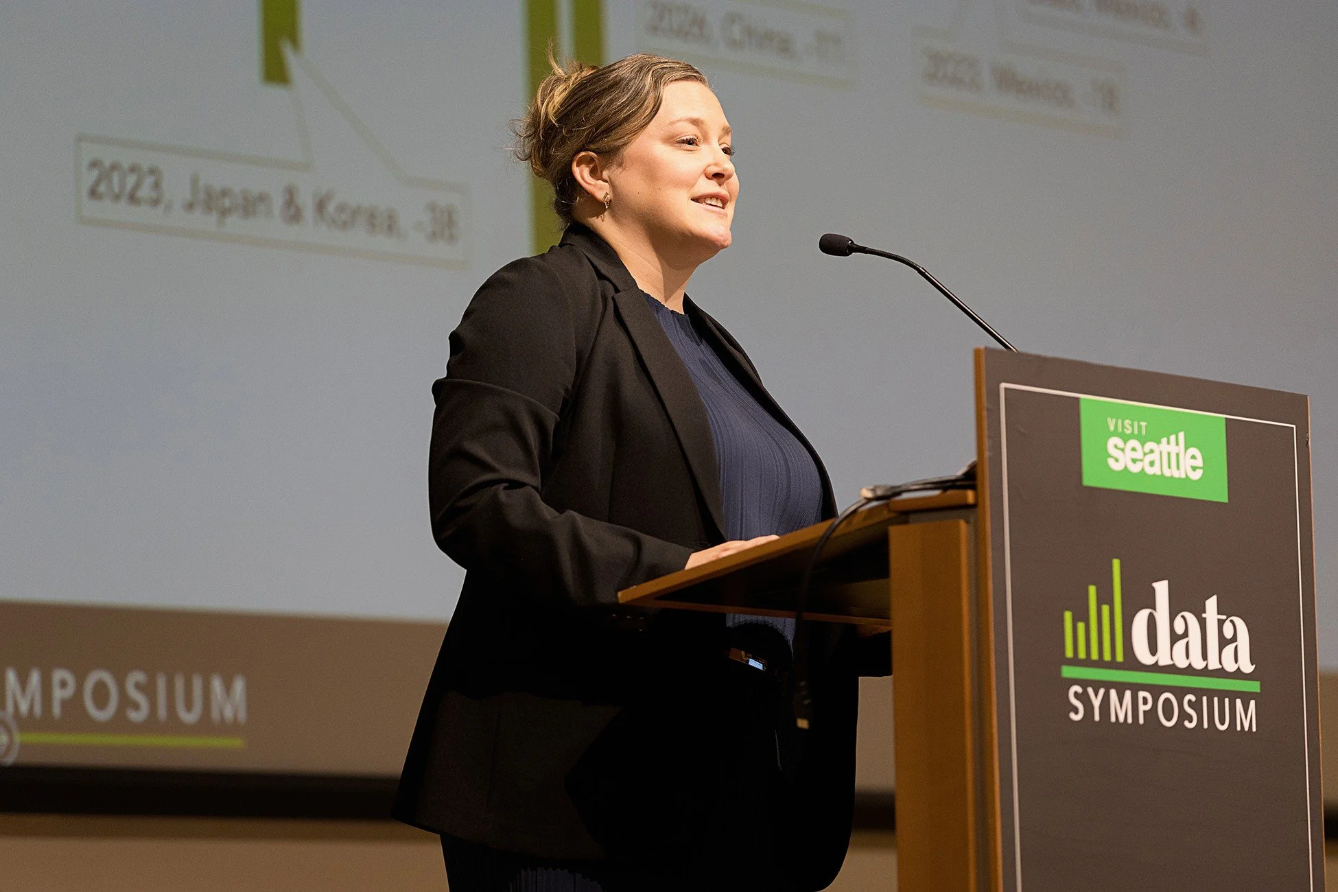 A woman speaking at a conference podium with a sign that reads 'data conference' and a screen in the background displaying a graph and the words '2023, Japan & Korea'.