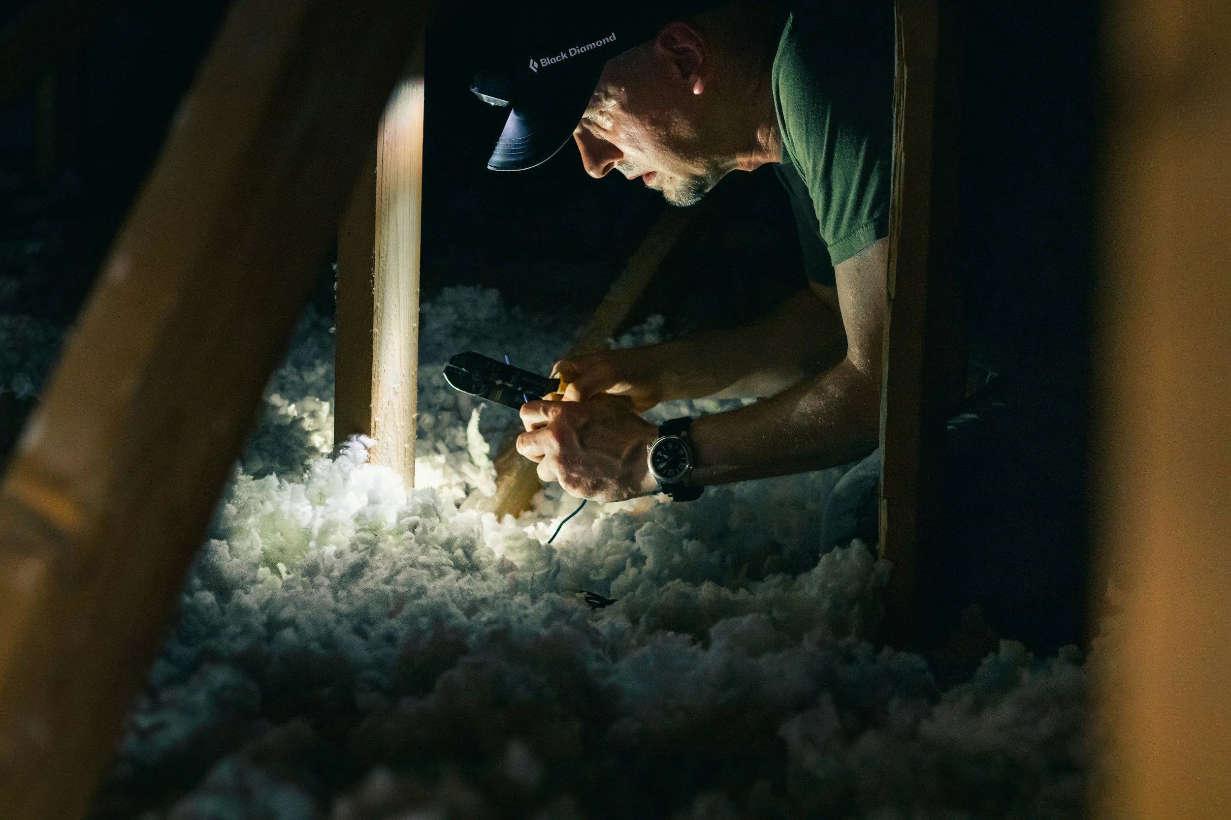 A man wearing a black cap and a watch is working with a tool in a confined space with wooden framing, surrounded by snow.