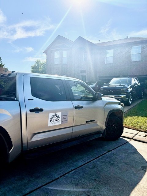 A silver pickup truck is parked in a driveway in front of a two-story brick house with a sunny sky. The truck has a sign on the door that says 'PEAK'.