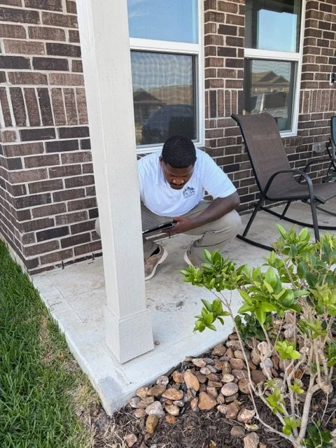 Man crouching on a patio next to a house wall, looking at his phone, with a chair and small bush nearby.
