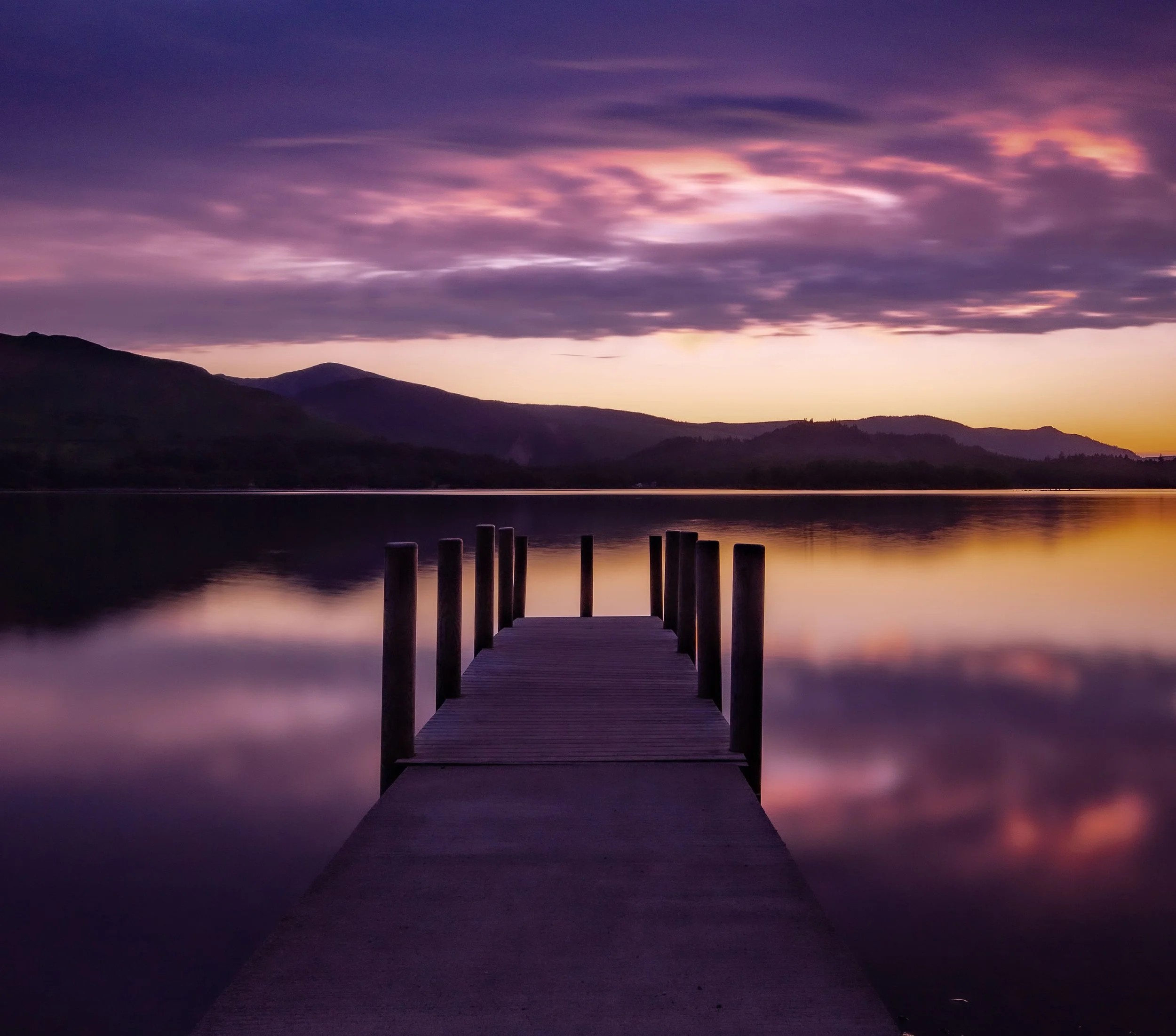 A wooden dock extends into a calm lake during sunset, with silhouettes of mountains and colorful purple, pink, and orange sky clouds reflected on the water.