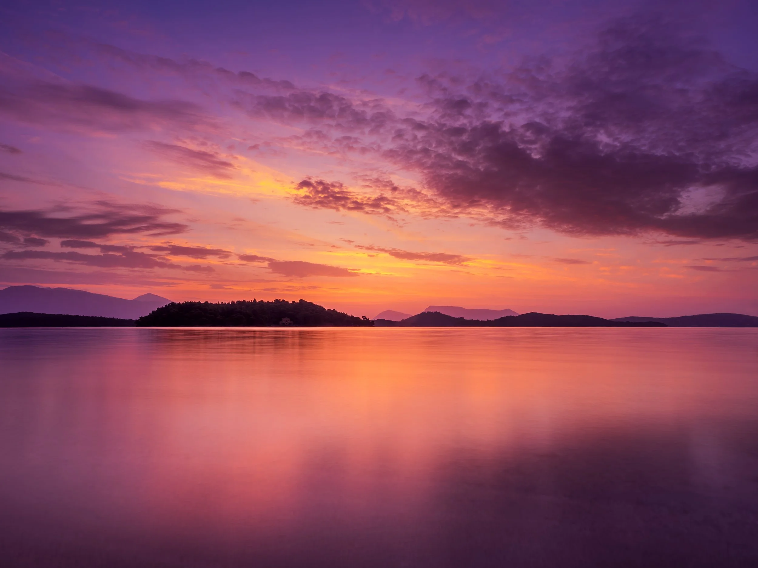 A vibrant sunset over a lake with silhouetted islands and mountains in the background, displaying shades of purple, pink, orange, and yellow in the sky.
