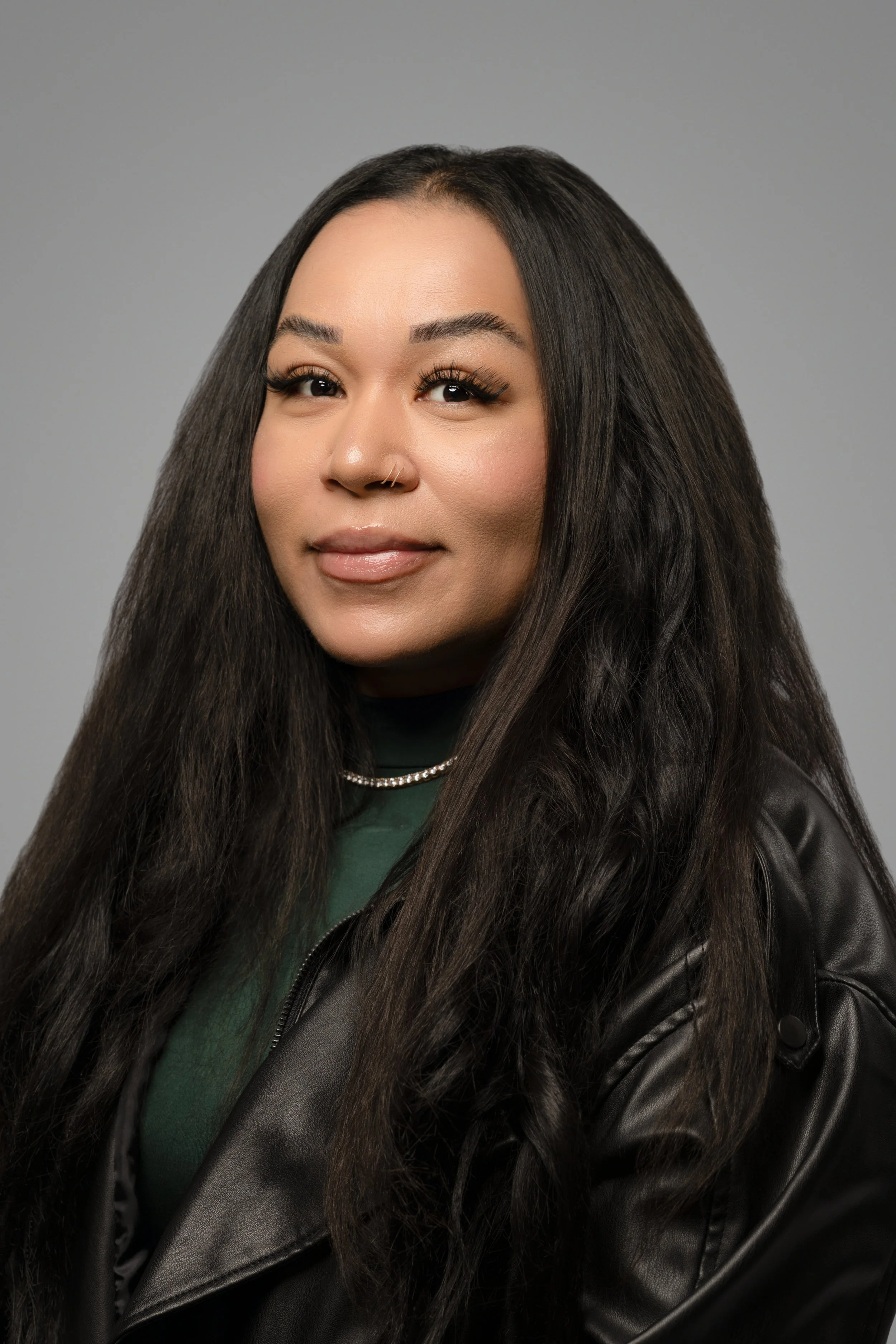 A woman with dark hair, wearing a black turtleneck and a red jacket, smiles at the camera, standing against a gray background.