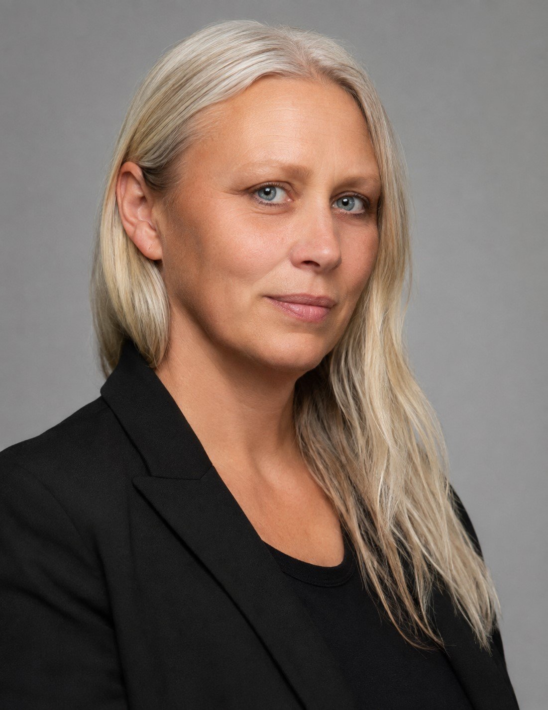 A woman with long, wavy, platinum blonde hair and blue eyes, wearing a black blazer and black top, standing against a plain gray background.