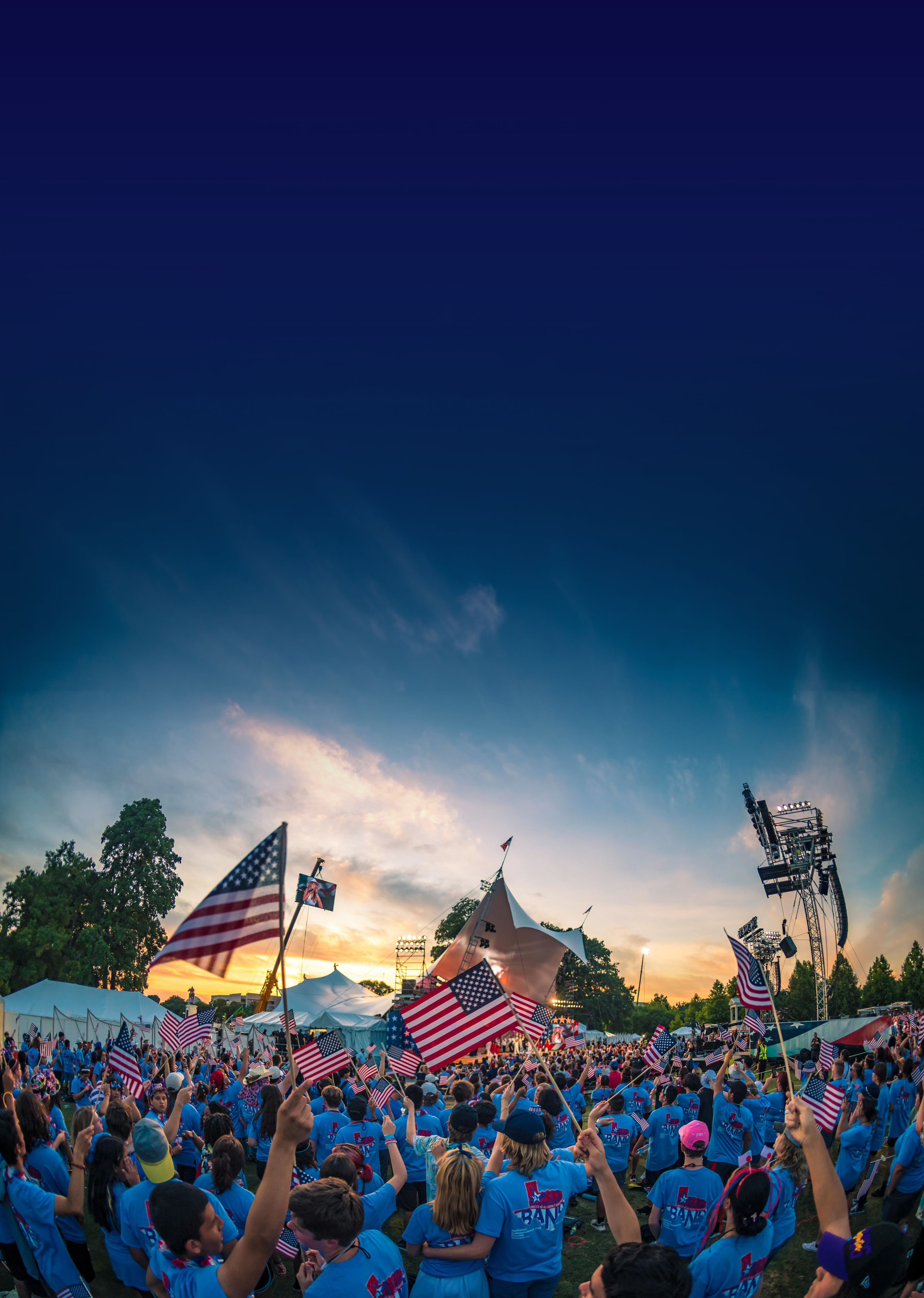 Crowd of people wearing blue shirts, holding American flags, gathered at an outdoor event during sunset. There are tents, stage equipment, and trees in the background.