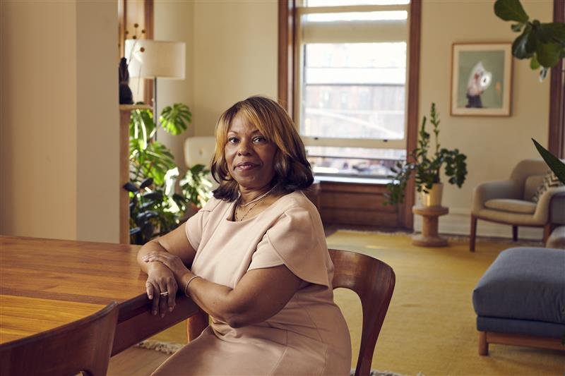 A woman sitting at a wooden table in a cozy, well-lit living room with large windows, indoor plants, and framed artwork.