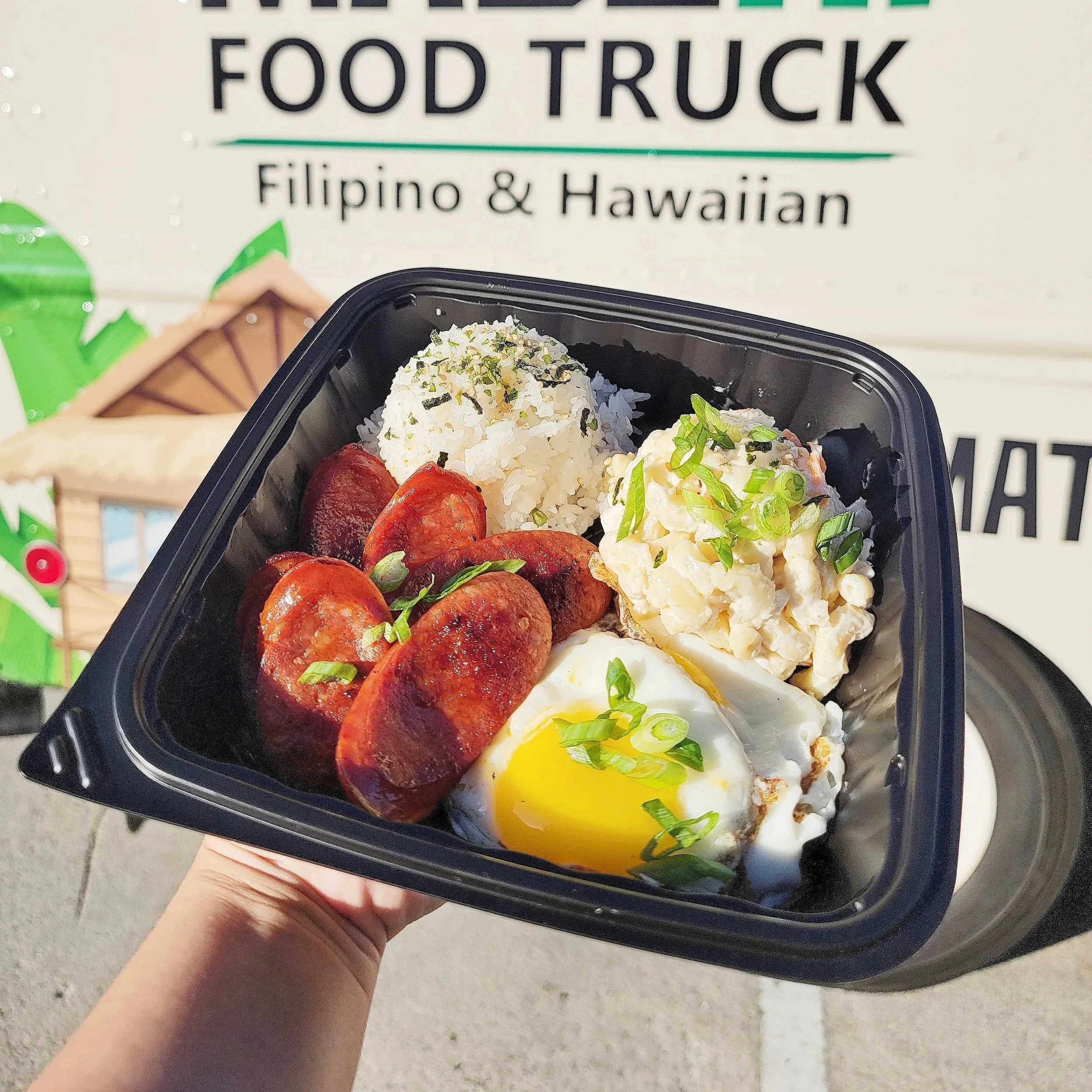 A person holding a black food container with assorted Hawaiian and Filipino foods in front of a sign that reads "Food Truck Filipino & Hawaiian".