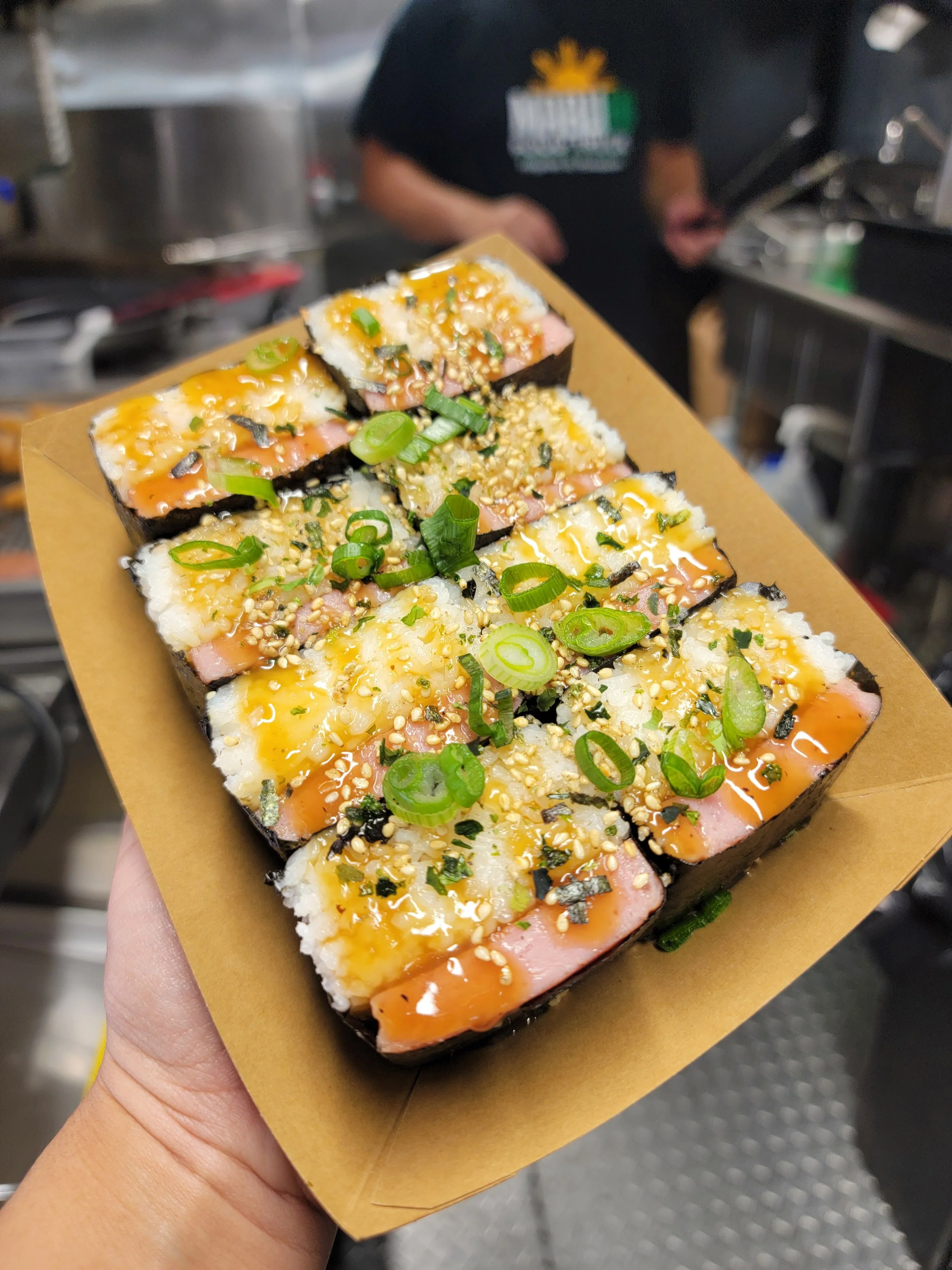 Hand holding a piece of sushi topped with green onions, sesame seeds, and sauce, with a blurred kitchen background.