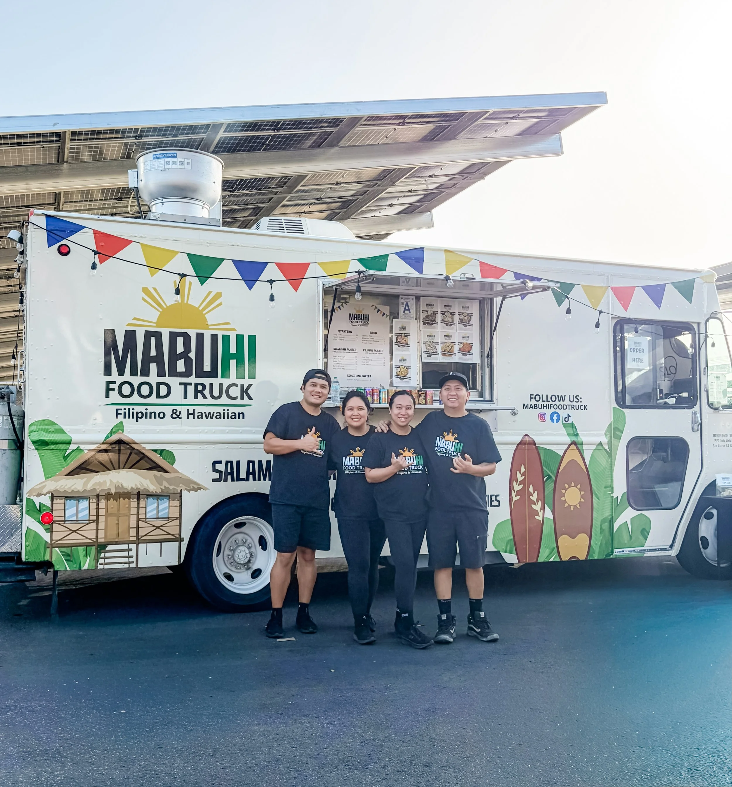 Four people standing in front of a food truck named Mabuhi Food Truck, decorated with colorful flags and tropical graphics, sunny outdoor setting.
