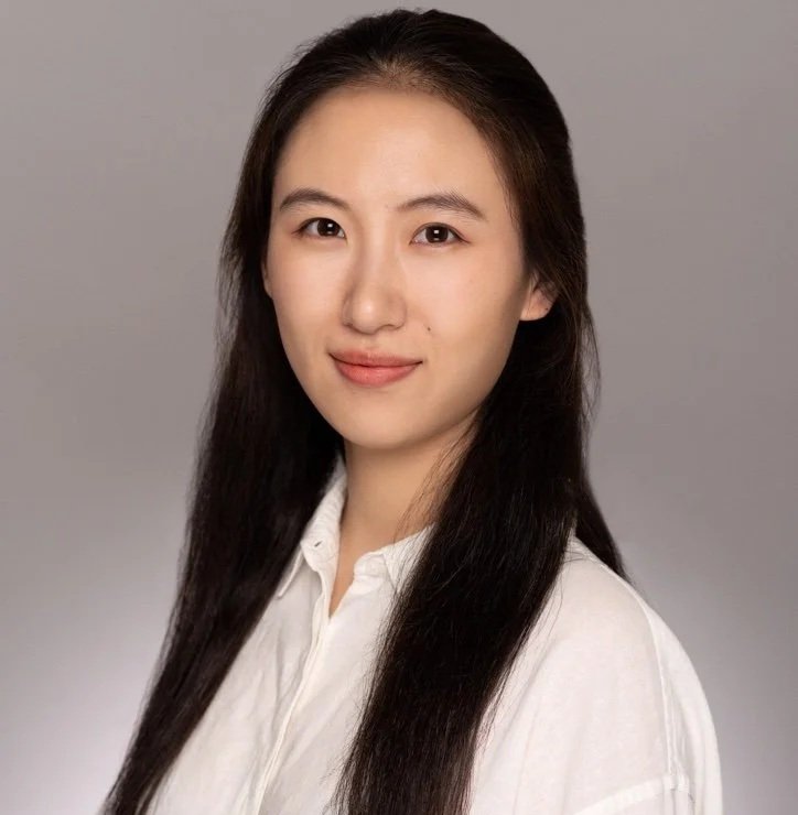 A young woman with long dark hair wearing a white shirt, looking at the camera and smiling slightly against a gray background.