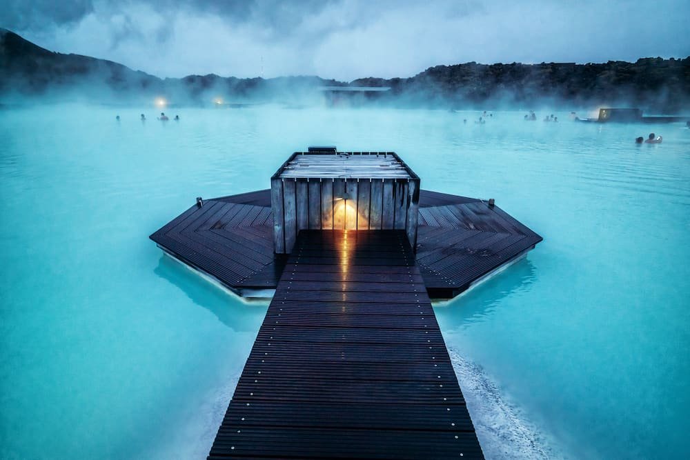 Wooden dock extending over hot spring with steam rising, surrounded by turquoise water, with people enjoying the bath in the distance, mountains in the background.