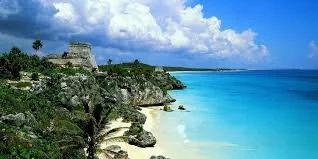 Tropical beachfront with white sand, turquoise water, and greenery on rocky cliffs under a partly cloudy sky.