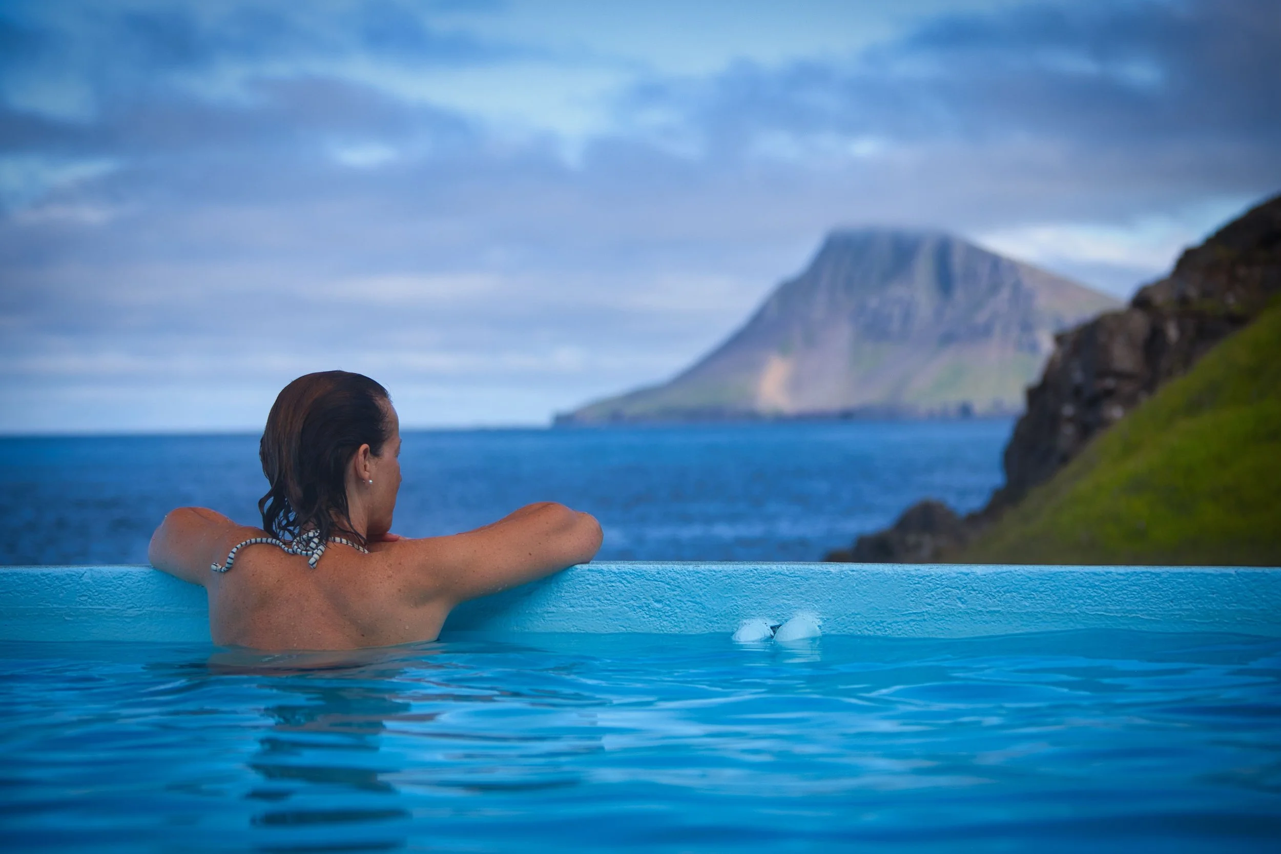 Back view of a woman relaxing in an outdoor thermal pool with scenic Icelandic mountains.