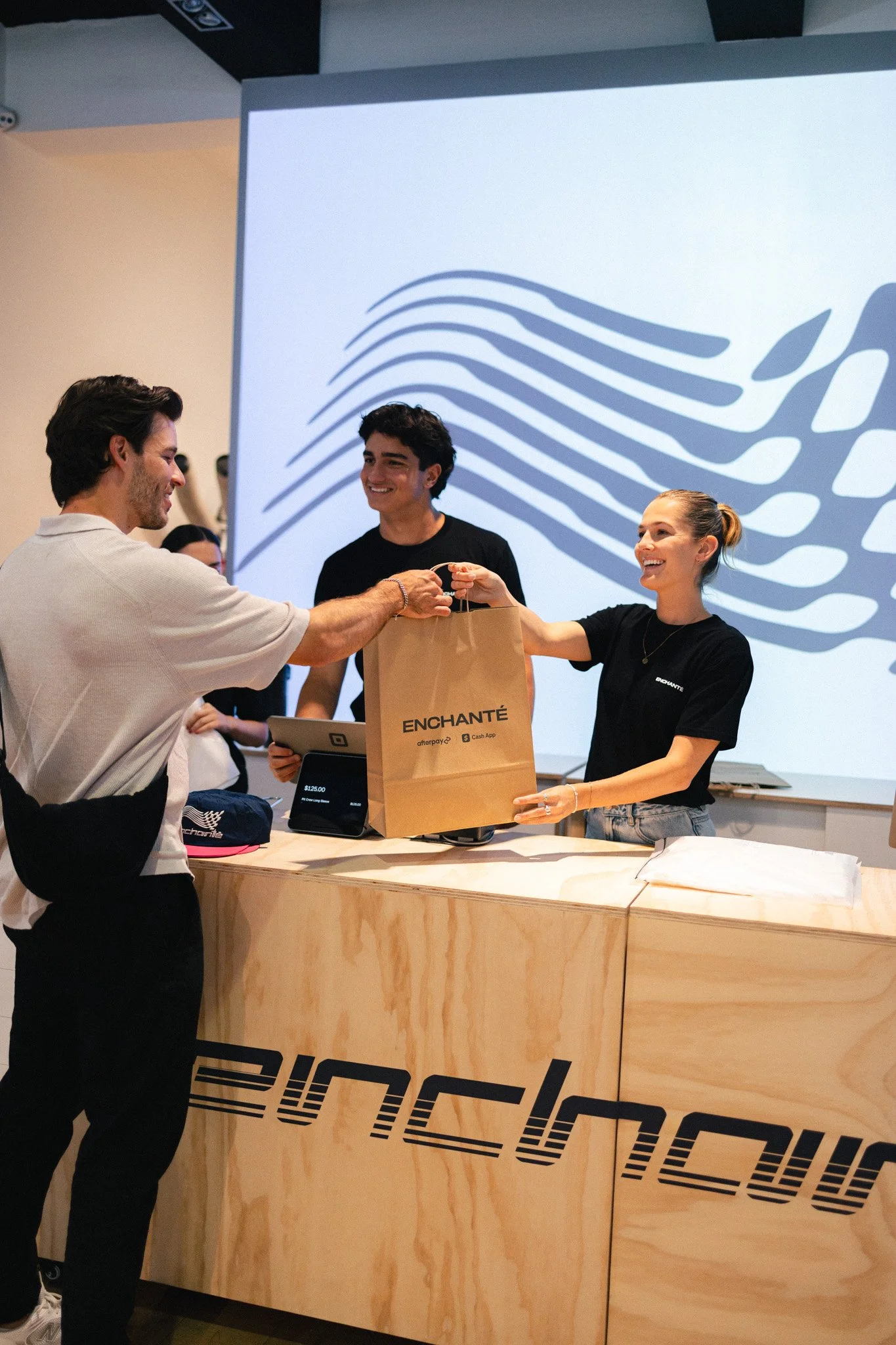 A man and a woman are standing at a wooden counter, exchanging a brown paper bag. The woman is smiling and wearing a black t-shirt, while the man is smiling and wearing a cream-colored shirt. There is a display screen and other people in the backgrou