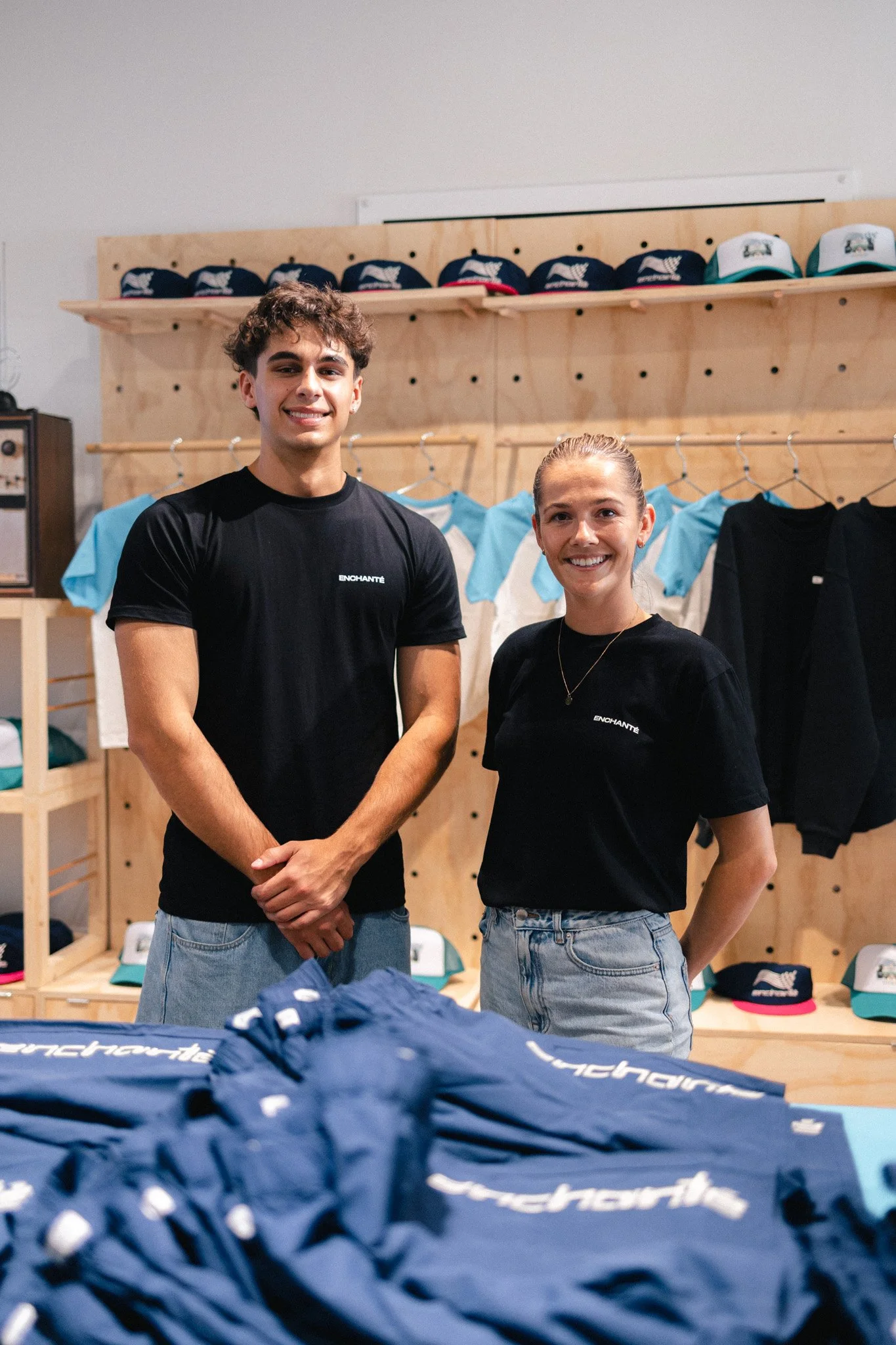 Two young adults, a man and a woman, standing inside a clothing store with wooden shelves and racks. They are smiling at the camera, and there are caps and folded clothes visible behind them. The foreground shows a table with navy blue clothing items