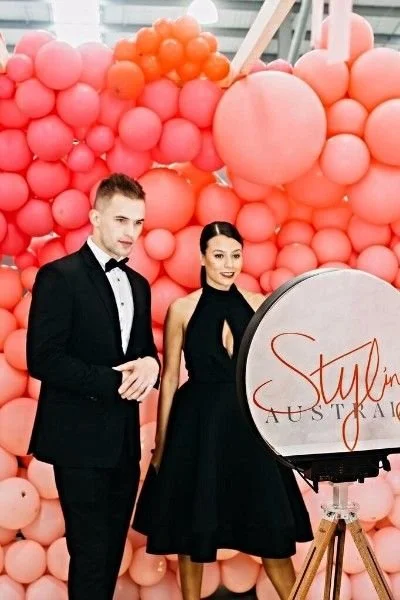 A man in a black tuxedo and a woman in a black dress standing in front of a pink balloon backdrop, with a sign that says "Stylen Australia."