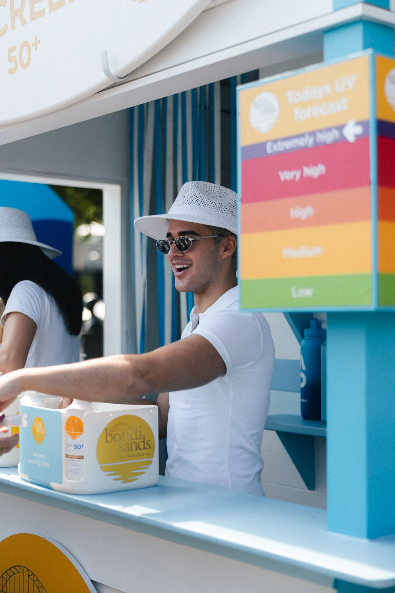 A young man with dark hair, wearing a white hat, sunglasses, and a white shirt, standing at a booth with a box of Bondi Sands sunscreen in front of him. Behind him, there is a colorful height or intensity scale sign and a blue and white themed setup.