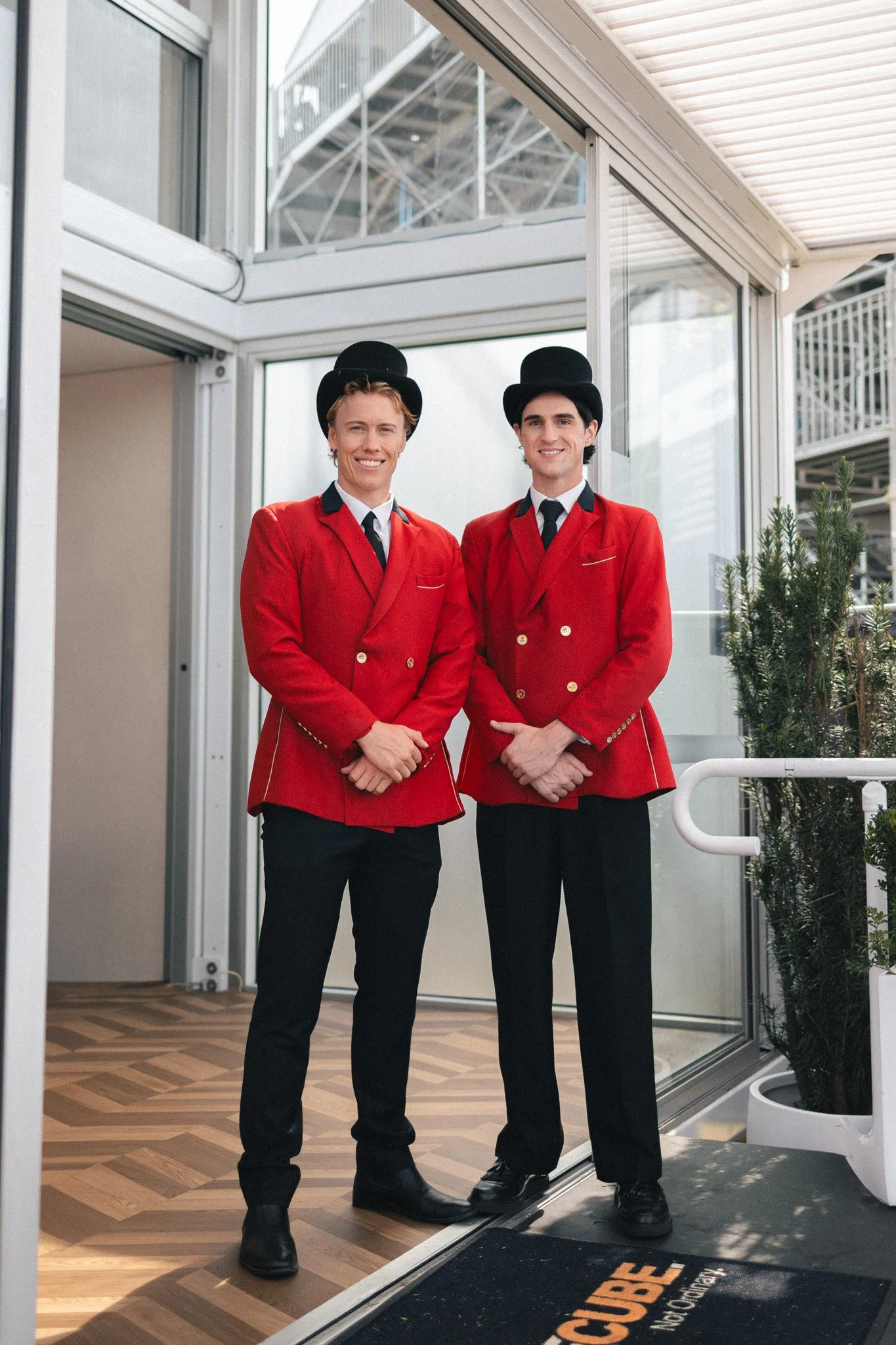 Two individuals dressed as hotel bellhops in red uniforms and black hats standing in the doorway of a building, smiling.