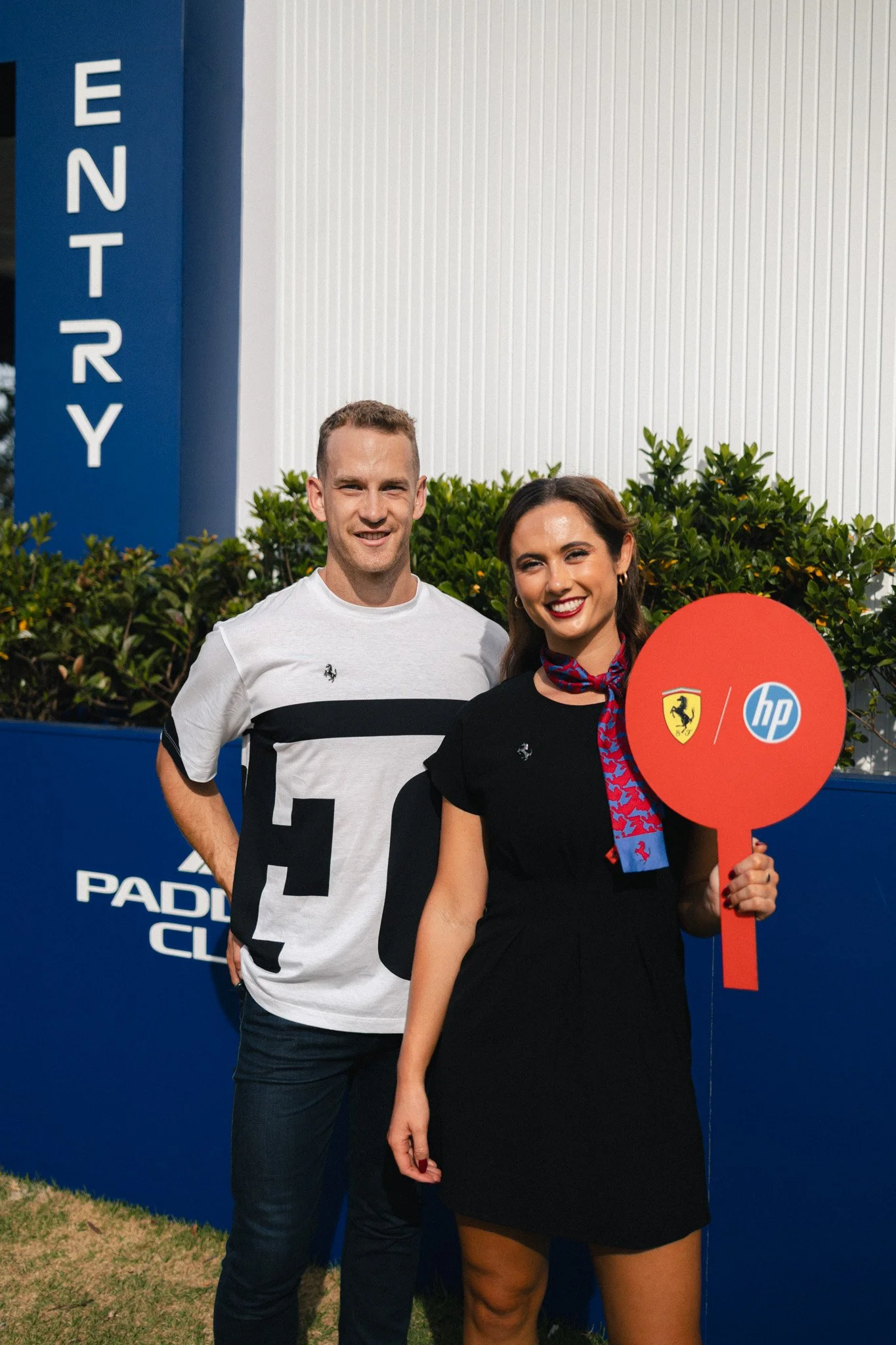 A man and a woman standing side by side outdoors in front of a blue wall with the word 'ENTRY' visible. The woman is holding a red paddle with logos of Ferrariand is smiling at the camera. The man is smiling and wearing a white t-shirt with black des