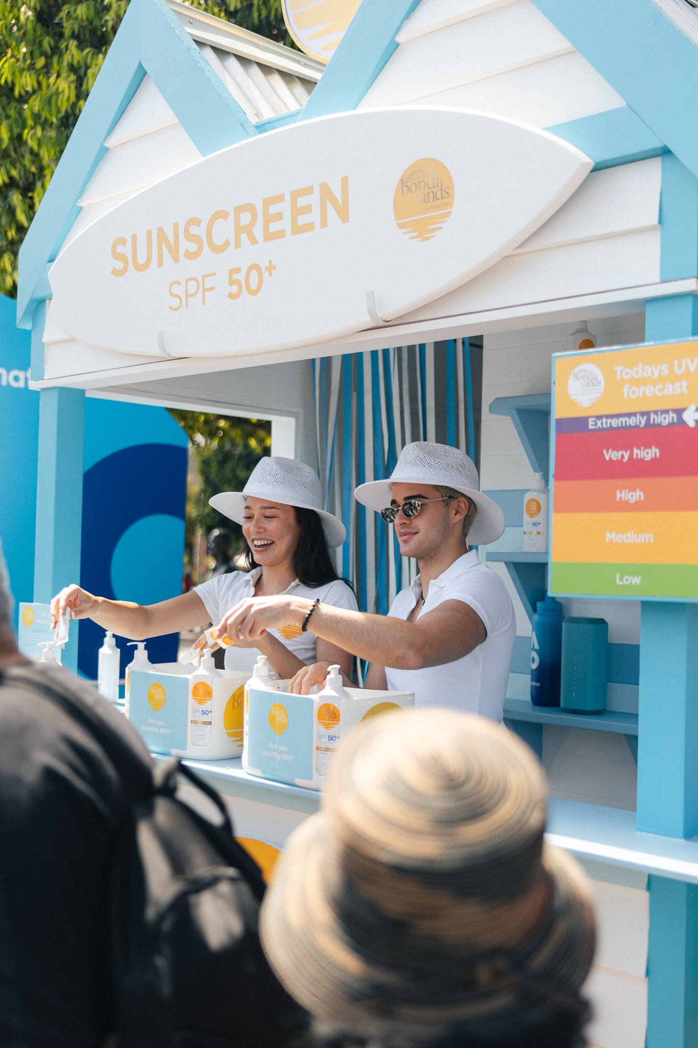 Two people working at a sunscreen booth with SPF 50+ label, crowd in front, sunny outdoor setting. Bondi Sands brand ambassadors sampling for the Australian Open - large scale sampling events