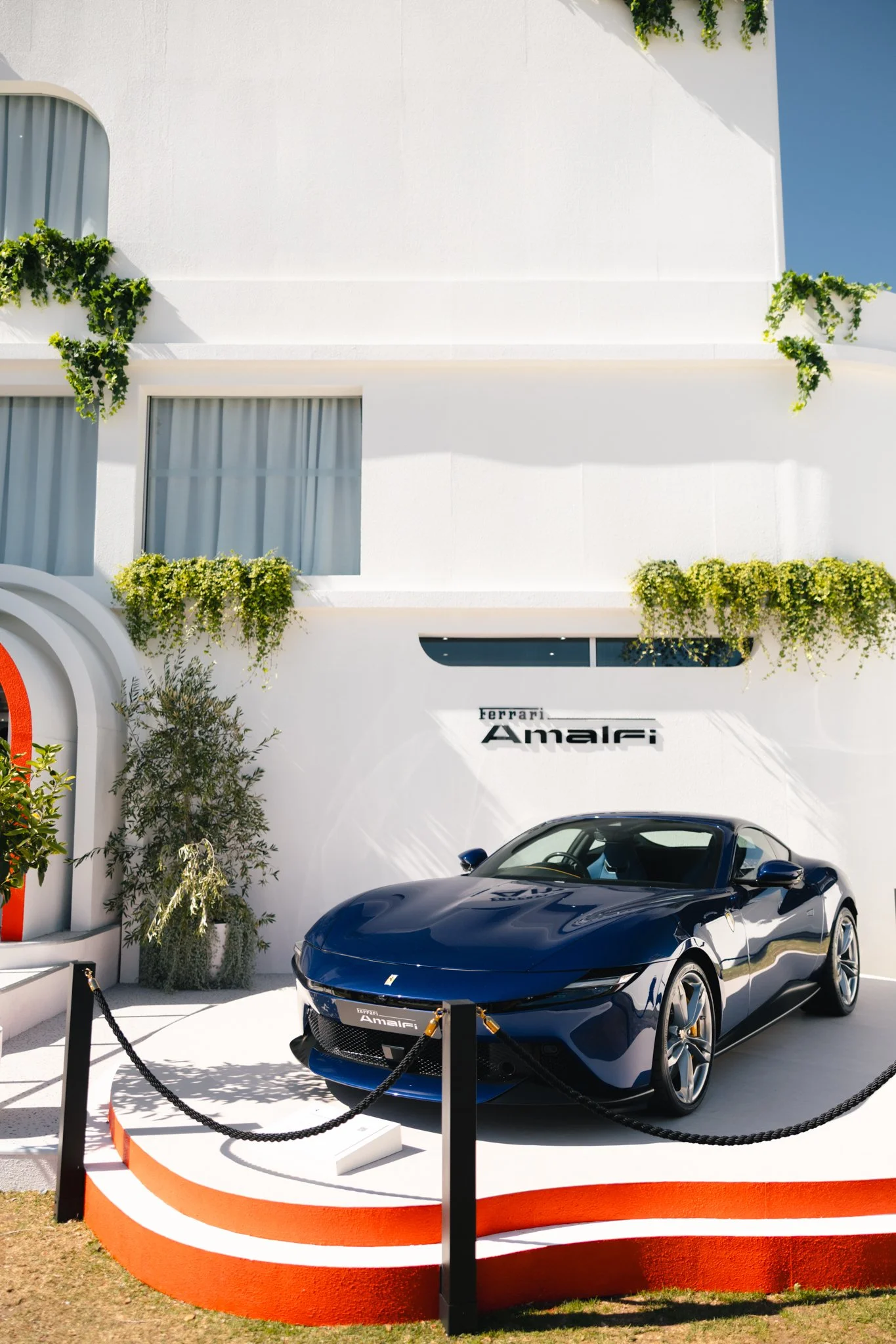A black Ferrari sports car on display outside a modern white building with green plants and blue sky in the background. The car is surrounded by black chain barriers with a sign that says 'Ferrari Amalfi'.